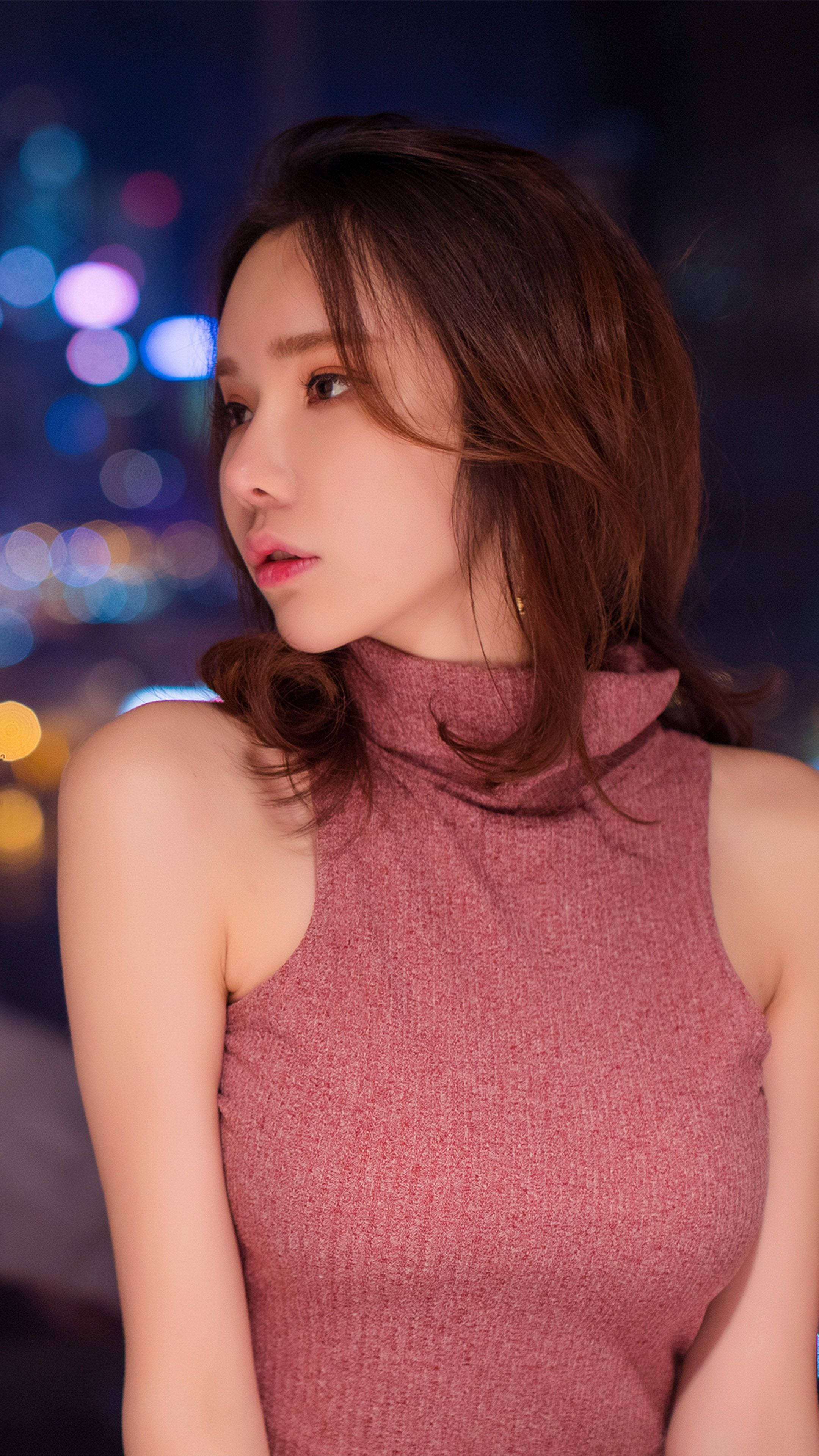 Cute Girl 桌布 壁紙 — 一位穿著粉紅色高領毛衣的女性，在充滿活力的散景城市夜景背景下若有所思地凝視著。