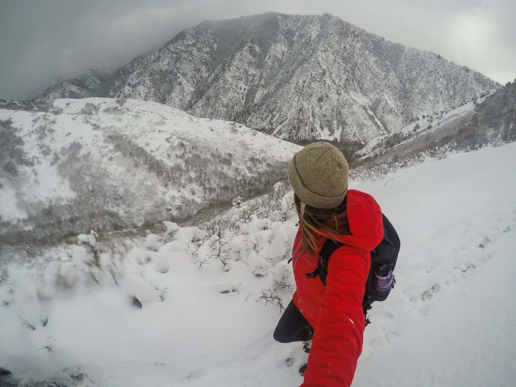 Winter Hiking 桌布 壁紙 — 一位穿著紅色外套和冷帽的遠足者在多雲的天空下穿越白雪覆蓋的山谷時自拍。