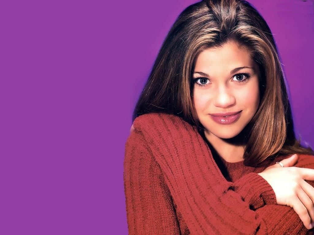 Danielle Fishel 桌布 壁紙 — 一位穿著紅色羅紋毛衣的年輕女性在純紫色攝影棚背景前輕柔微笑。