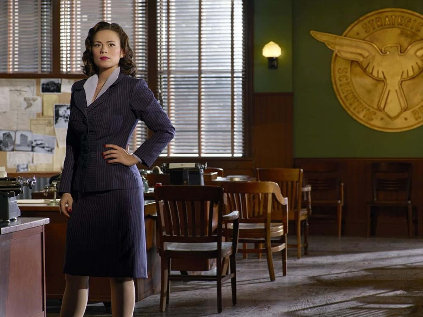 Peggy Carter 桌布 壁紙 — 一位穿著細條紋西裝的女性自信地站在一個配有木製家具和牆上大型老鷹標誌的復古辦公室中。