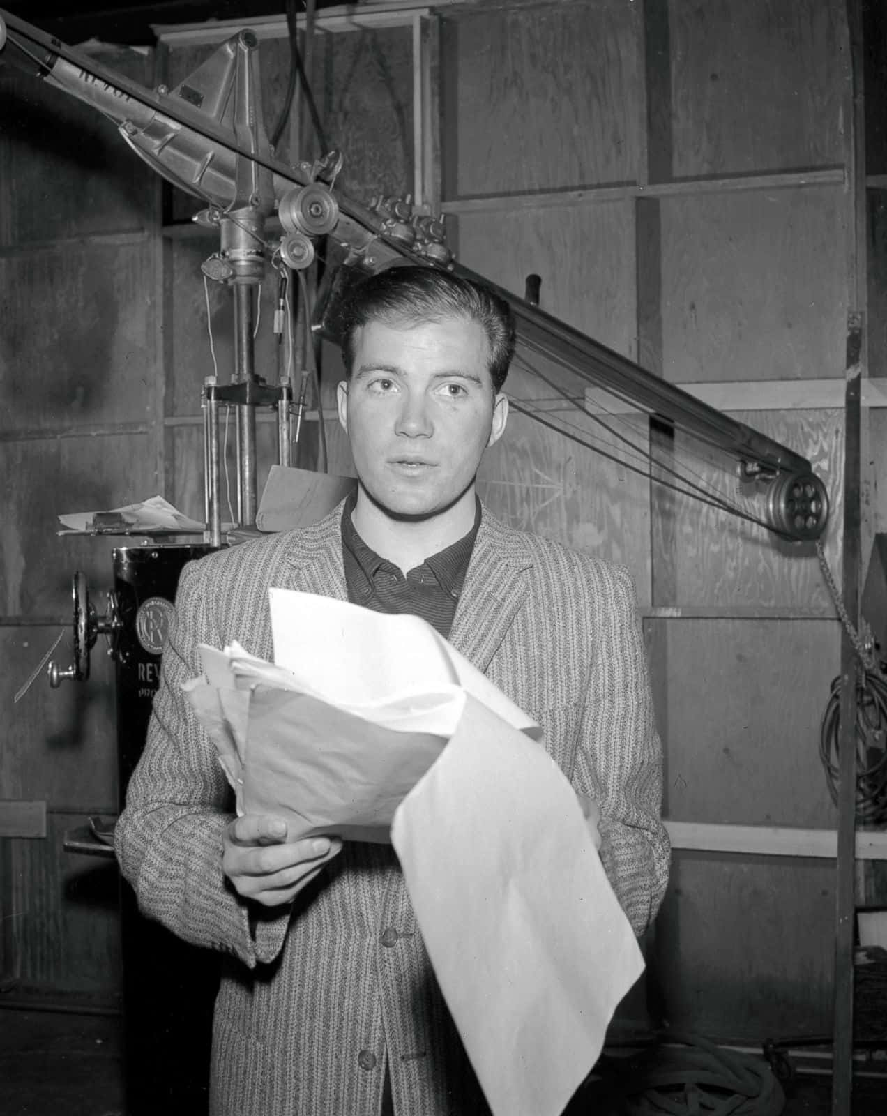 william shatner 桌布 壁紙 — 一位穿著細條紋西裝的年輕男子在復古電影製片廠中拿著文件，身後可見攝影器材。
