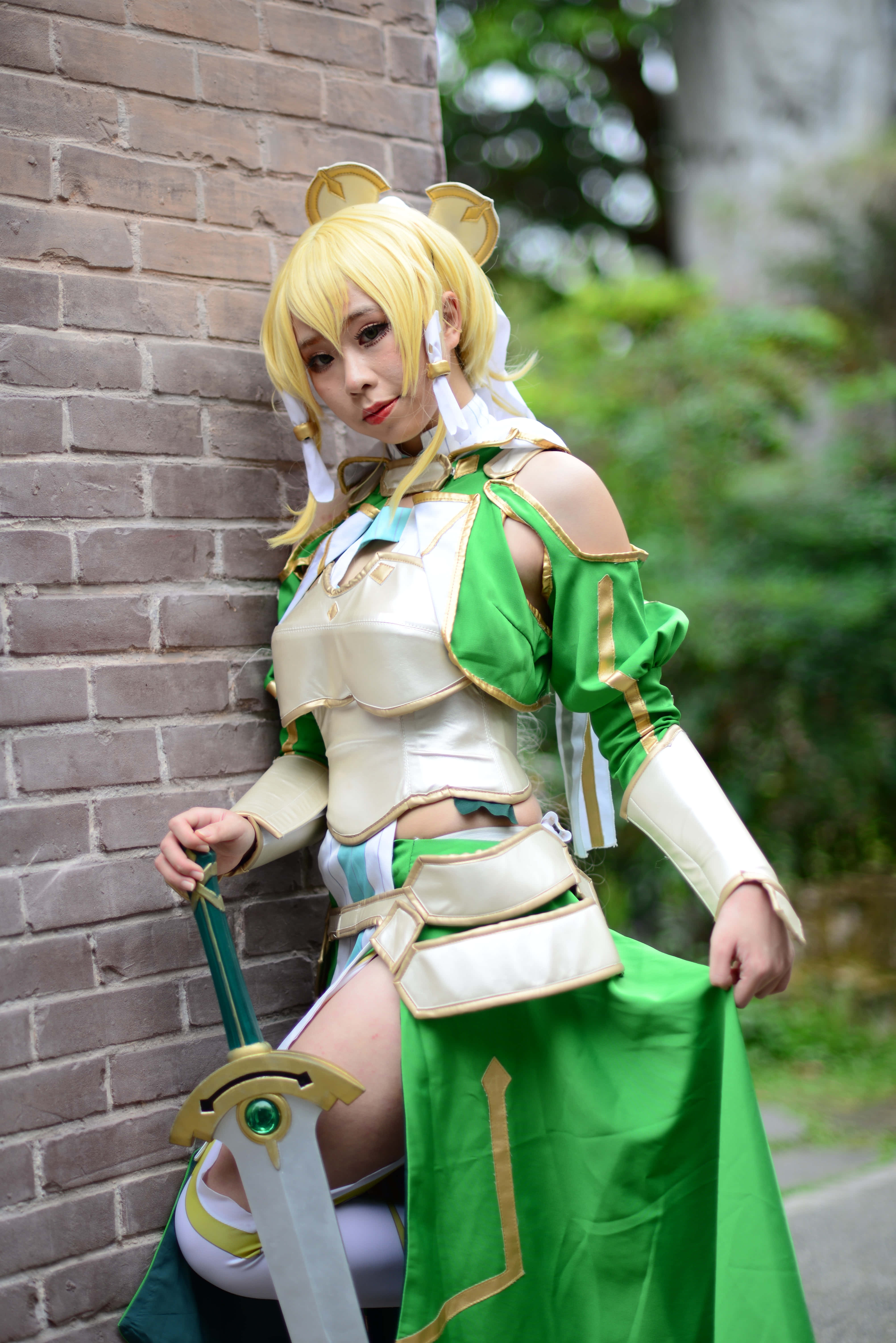 Sword Art Online Cosplay 桌布 壁紙 — 一位穿著綠色和奶油色奇幻盔甲的角色扮演者靠著磚牆，手持一把帶有綠色寶石的劍。