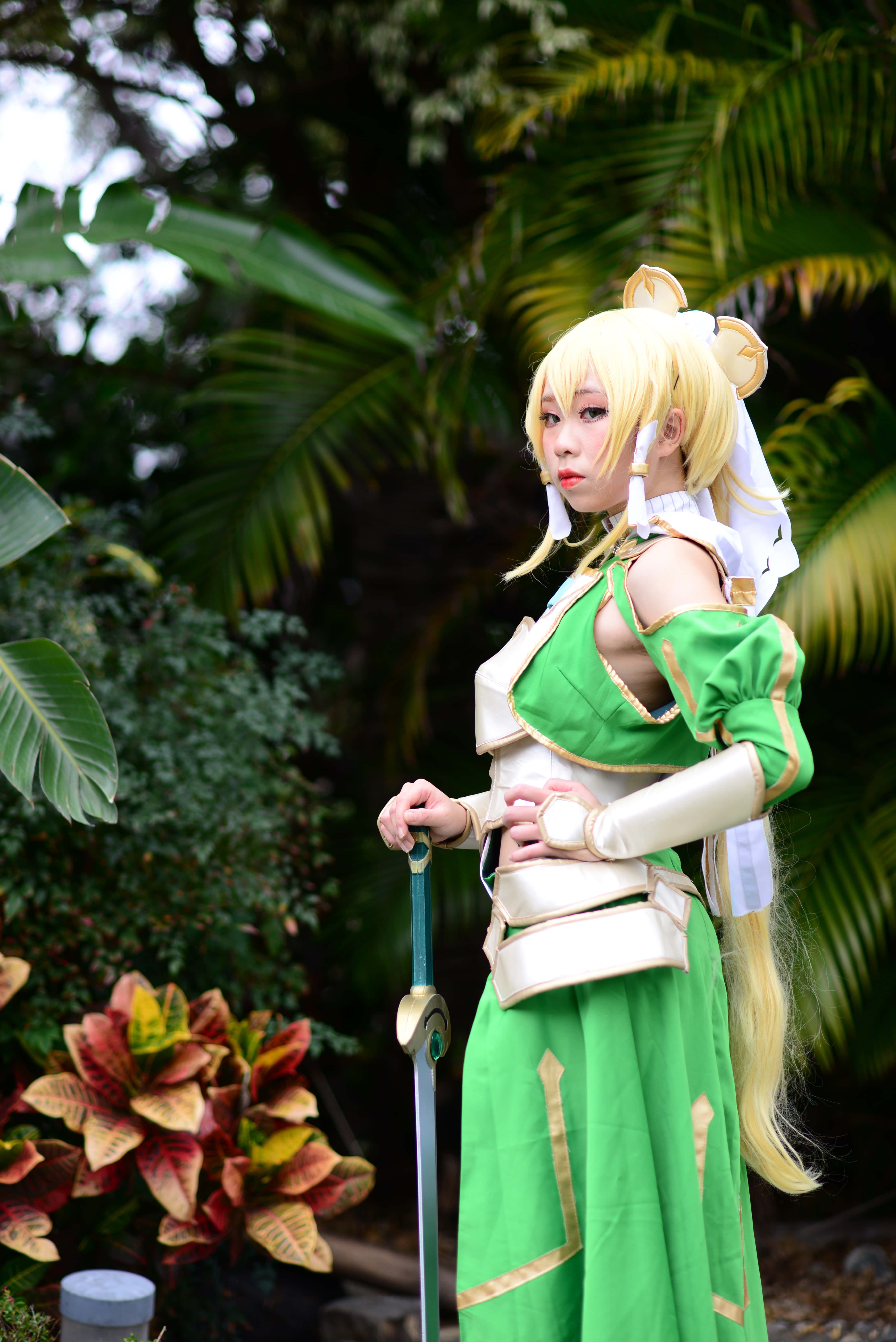 Sword Art Online Cosplay 桌布 壁紙 — 一位穿著綠色和白色奇幻服裝、戴著狐狸耳朵的角色扮演者在茂密的熱帶植物中手持一把劍。