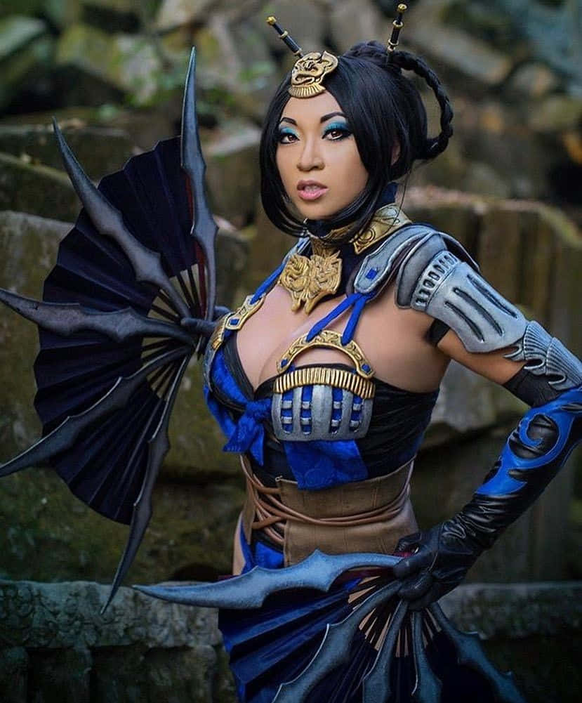 Mortal Kombat Kitana 桌布 壁紙 — 一位穿著華麗藍金色盔甲、手持尖刺扇狀武器的角色扮演者，在長滿青苔的石牆前自信地擺出姿勢。