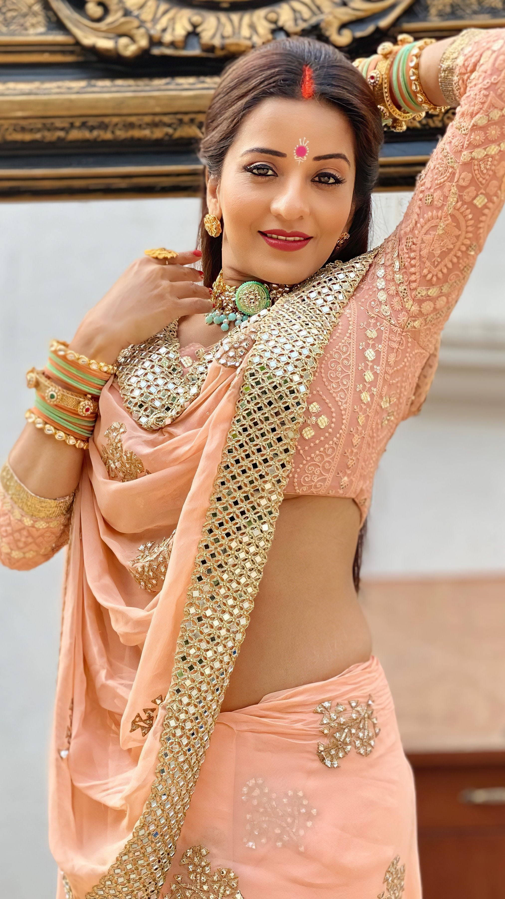 Bhojpuri 桌布 壁紙 — 一位穿著華麗蜜桃色和金色傳統服飾的女性，佩戴珠寶和眉心貼，在裝飾性背景前優雅地擺姿勢。