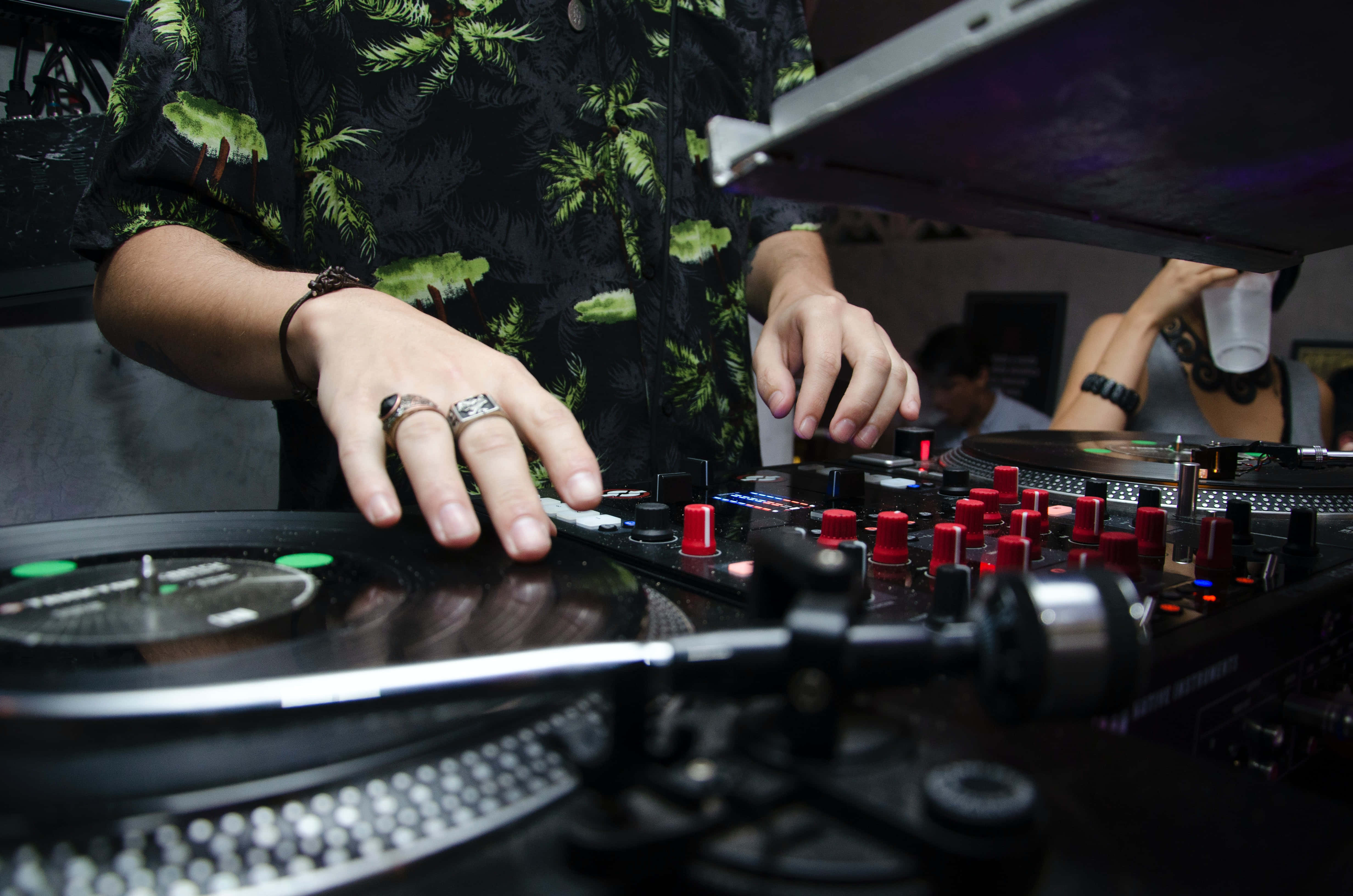 DJ 桌布 壁紙 — 一位穿著葉子圖案襯衫的DJ在昏暗的俱樂部燈光下，在帶有紅色旋鈕發光的唱盤機上混音唱片。