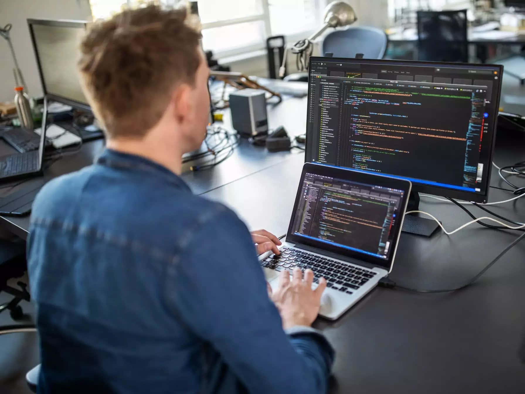 Programming 桌布 壁紙 — 一位穿著藍色襯衫的程式設計師在現代辦公空間中，在筆記型電腦和顯示器上輸入程式碼。