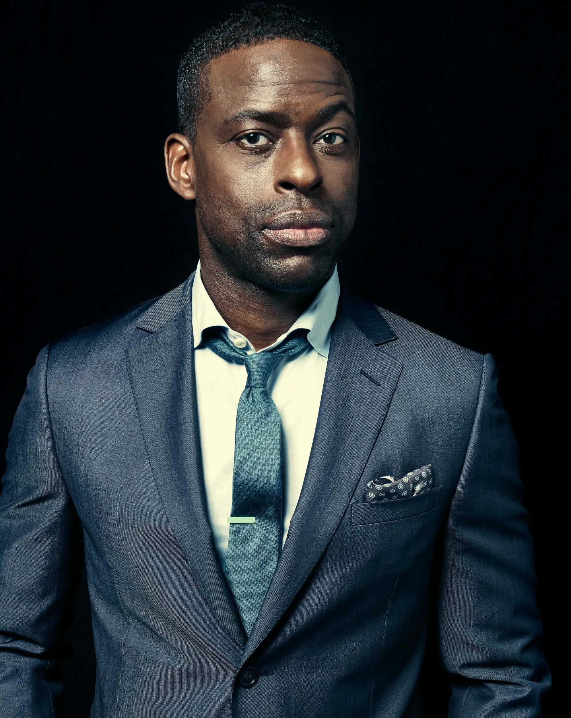 Sterling K Brown 桌布 壁紙 — 一位穿著藍色西裝和領帶、衣著講究的男子，在黑色背景前凝視著。