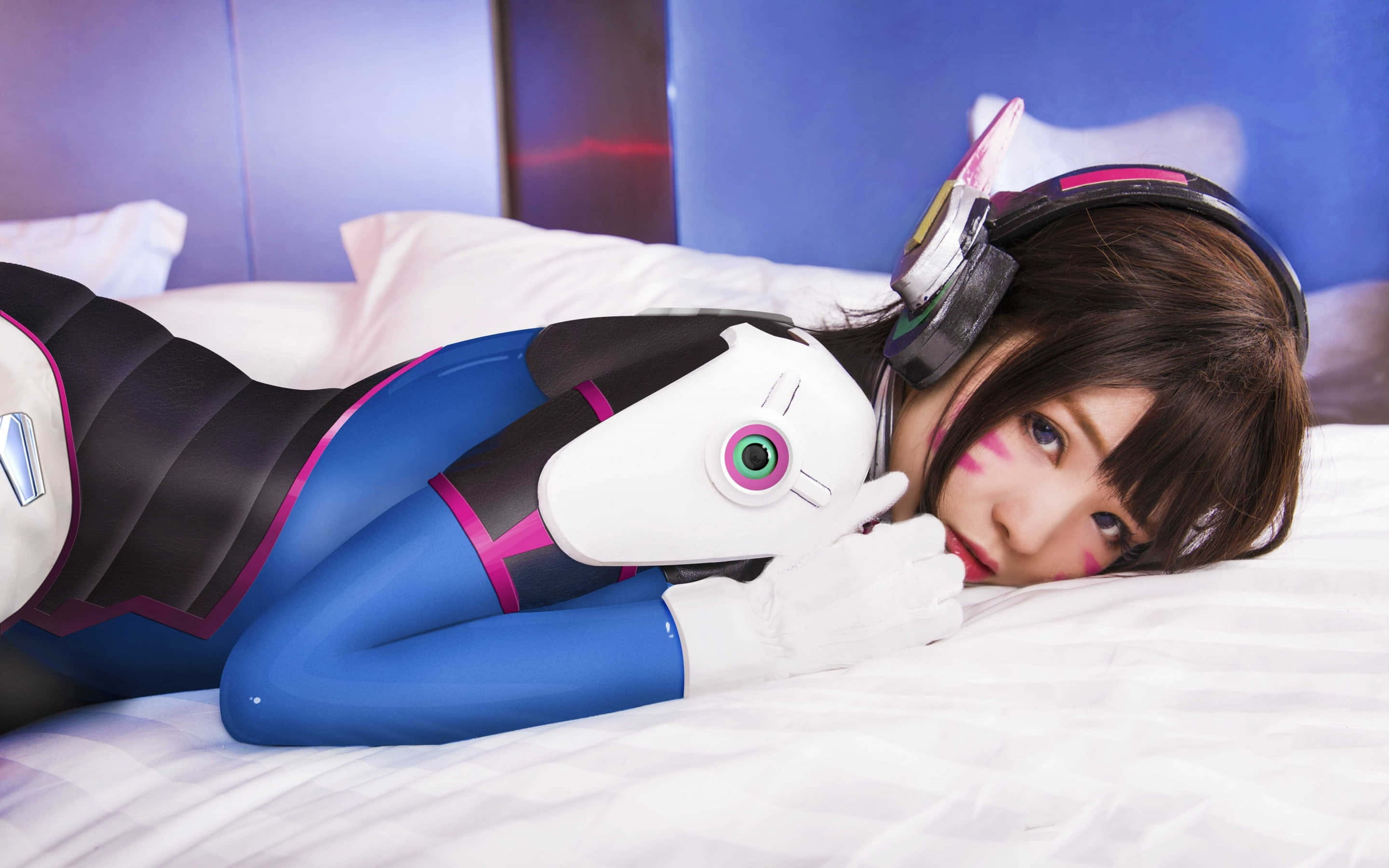 Gaming Cosplay 桌布 壁紙 — 一位穿著藍黑粉紅色Overwatch Tracer服裝的角色扮演者躺在床上，手持一個帶有綠色眼睛的白色頭盔。