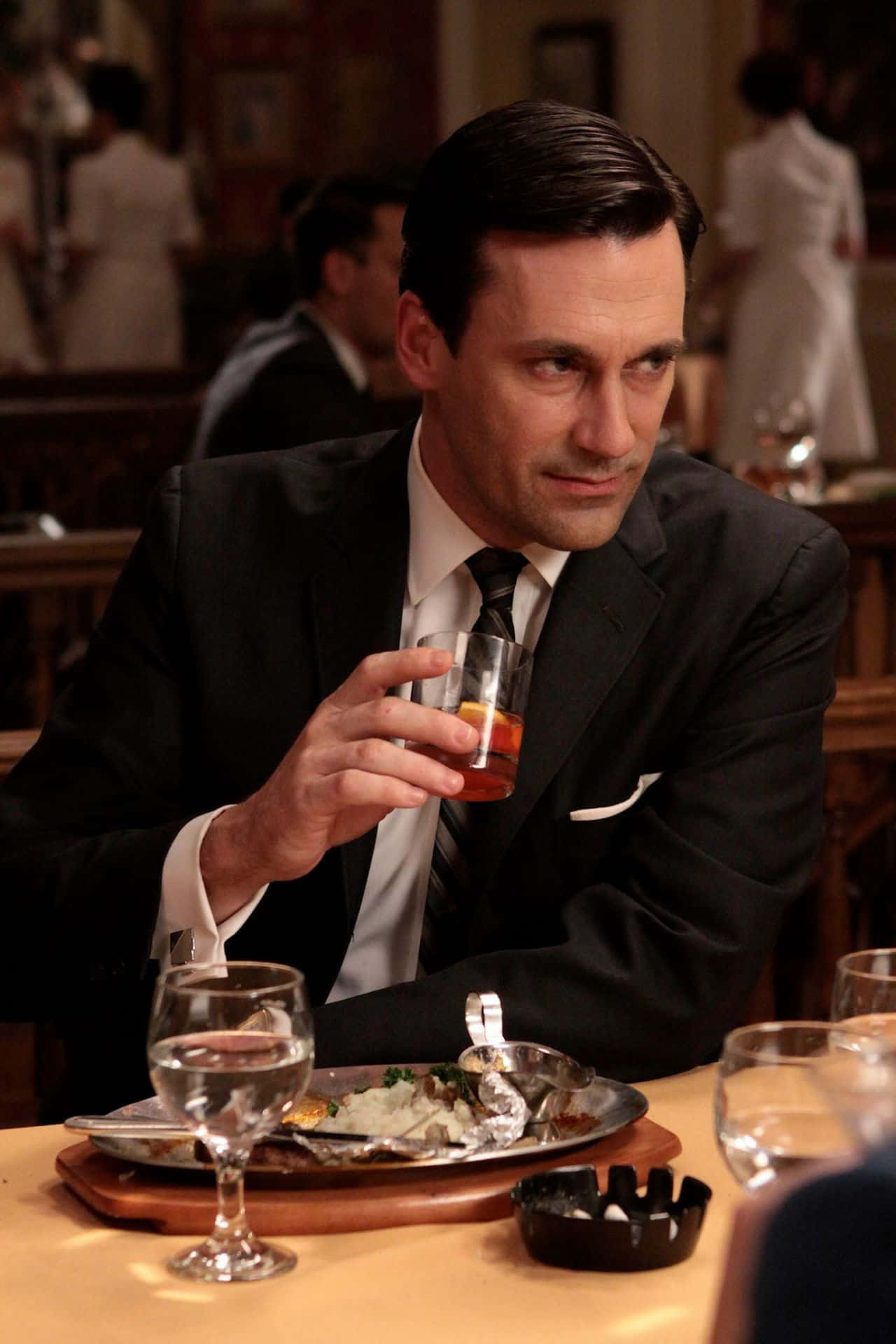 Jon Hamm 桌布 壁紙 — 一位穿著西裝、打扮俐落的男士在燈光昏暗的餐廳桌上，一邊享用食物和煙灰缸，一邊啜飲威士忌。