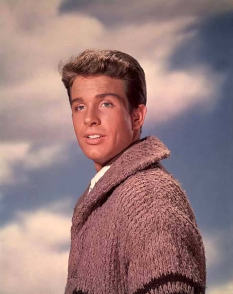Warren Beatty 桌布 壁紙 — 一位穿著質感針織開襟衫的年輕男子，在陰天背景下轉頭越過肩膀向後看。