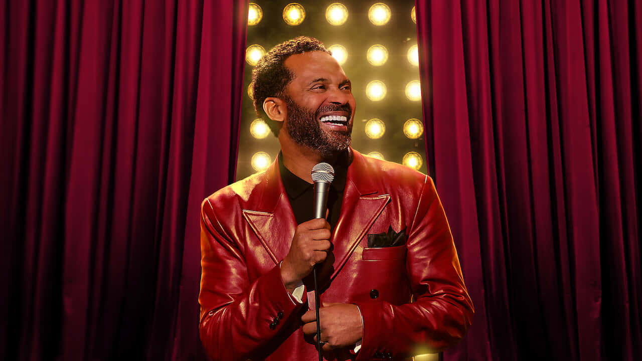 Mike Epps 桌布 壁紙 — 一位穿著閃亮紅色皮褸的喜劇演員笑著在舞台上拿著咪高峰，背景是栗色窗簾和閃爍的聚光燈。