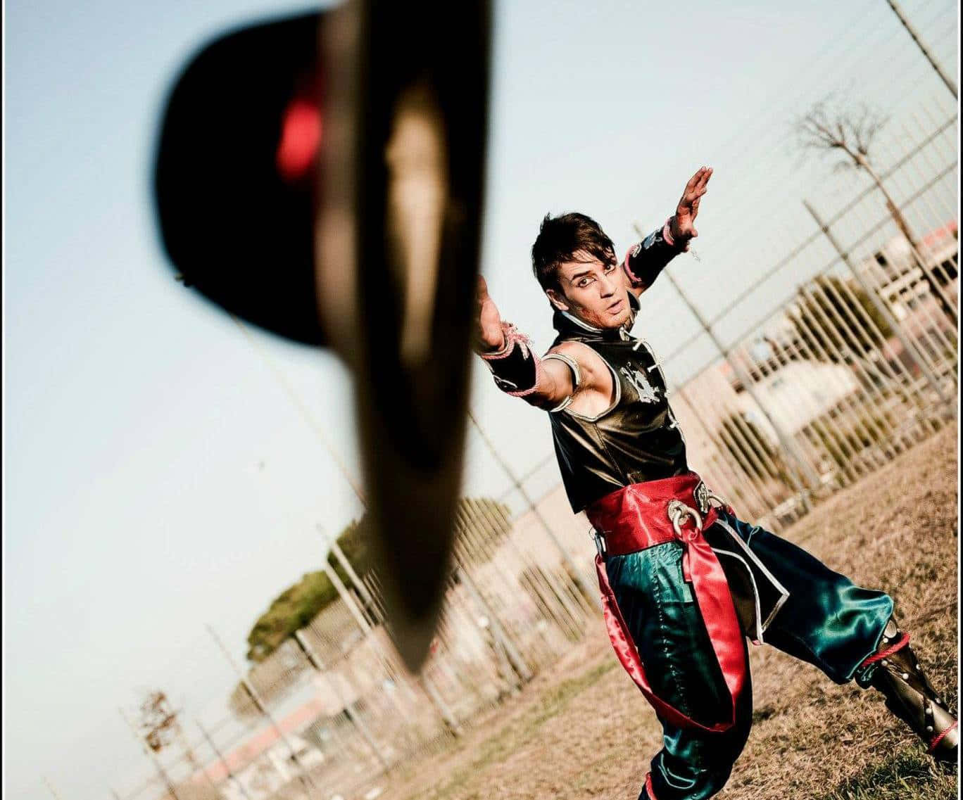 Mortal Kombat Kung Lao 桌布 壁紙 — 一位穿著閃亮黑色和紅色服裝的角色扮演者在戶外擺出動態武術姿勢，前景有一個模糊的圓形物體。