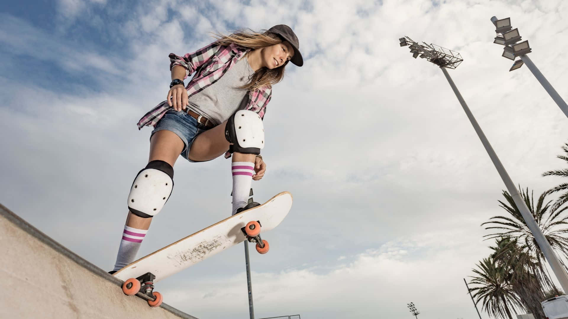 滑板 桌布 壁紙 — 一位穿著防護裝備的年輕女性在陰天背景下，在斜坡上玩滑板，背景有棕櫚樹和體育場燈光。