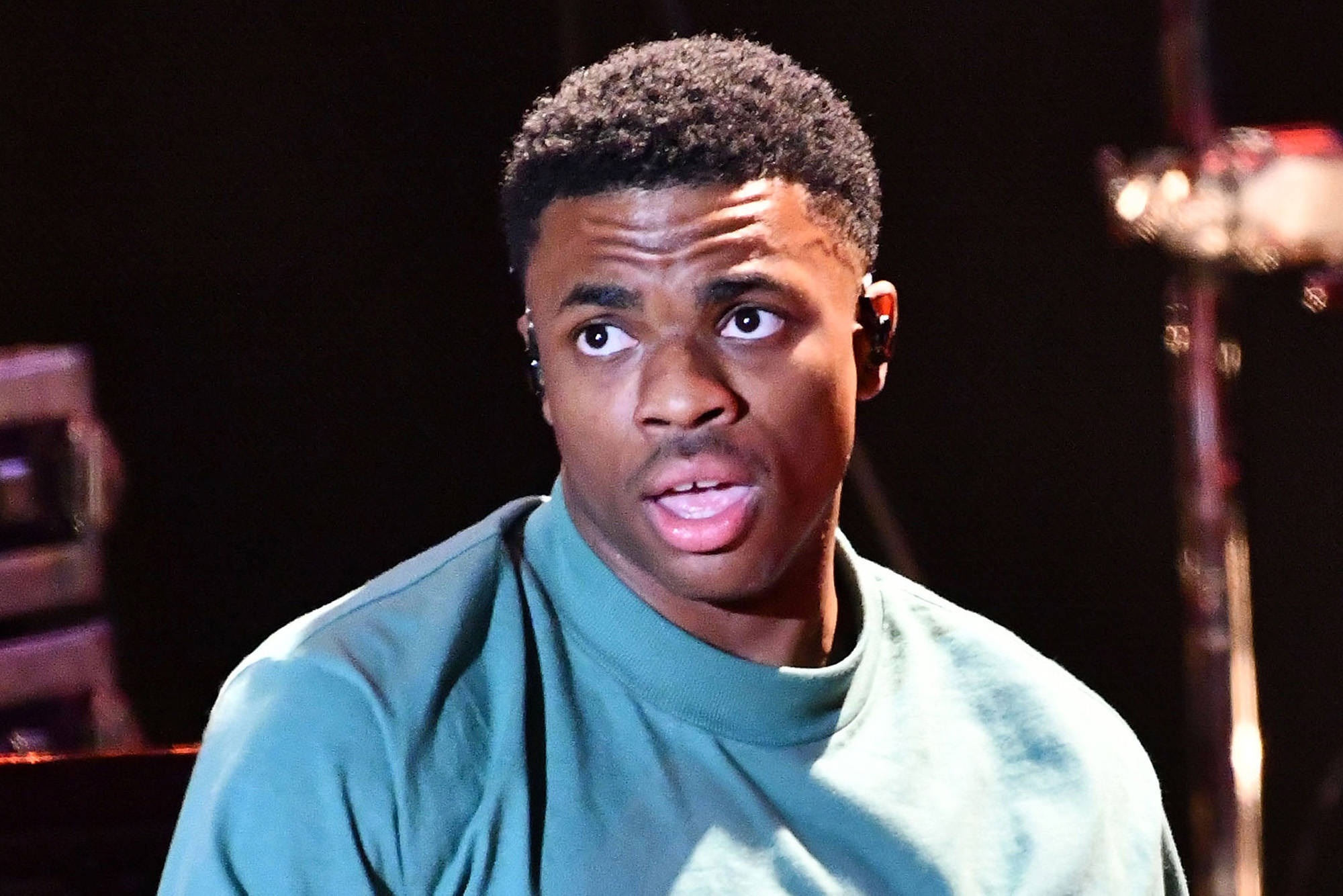 Cutthroat Boyz Vince Staples 壁紙 — 一位穿著青綠色高領毛衣的年輕黑人男子在昏暗的舞台上，背景有爵士鼓，表情驚訝。
