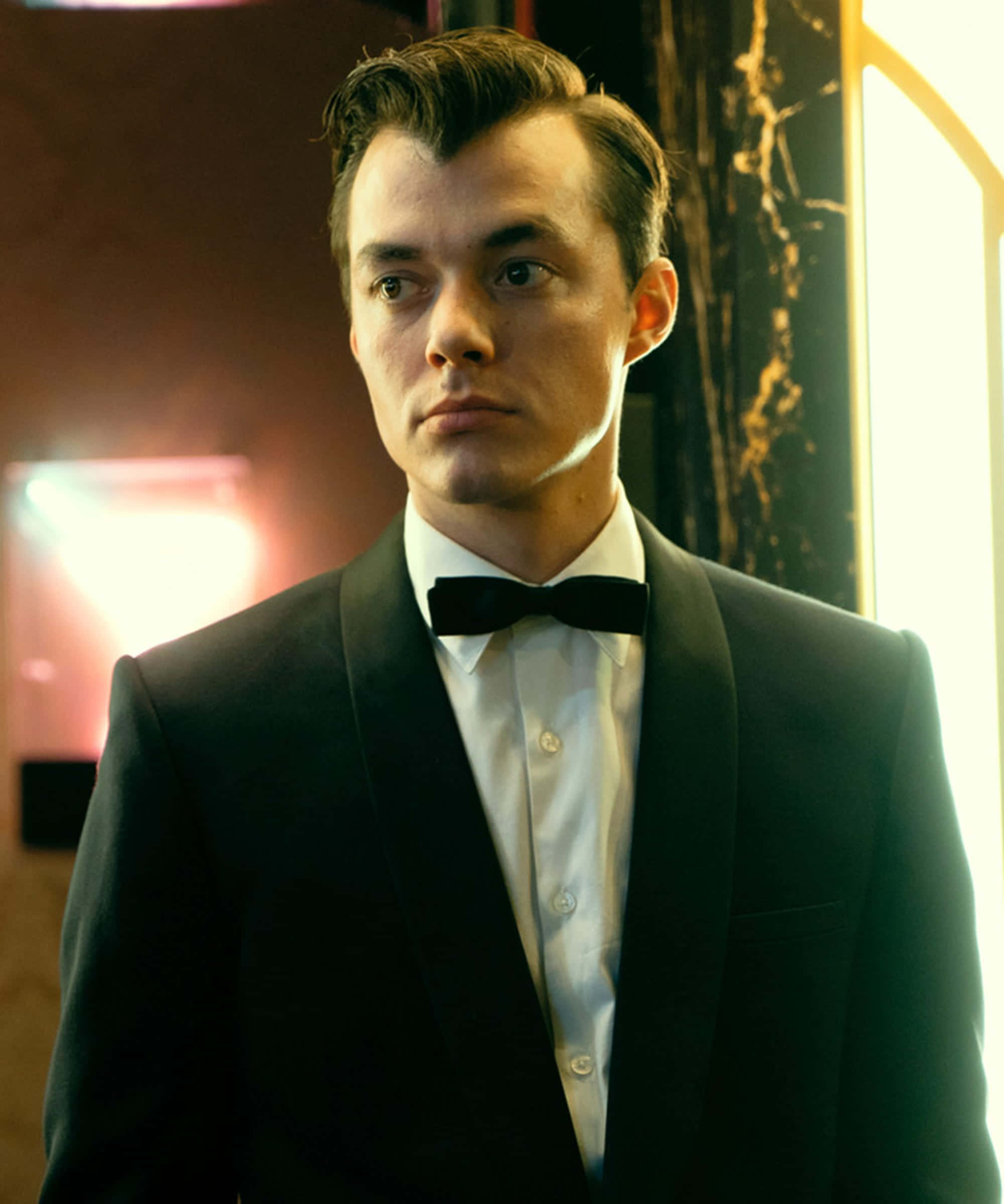 Alfred Pennyworth 桌布 壁紙 — 一位穿著黑色燕尾服和領結、衣著講究的男士，站在一個光線昏暗、帶有溫暖環境照明和大理石牆壁的房間裡。