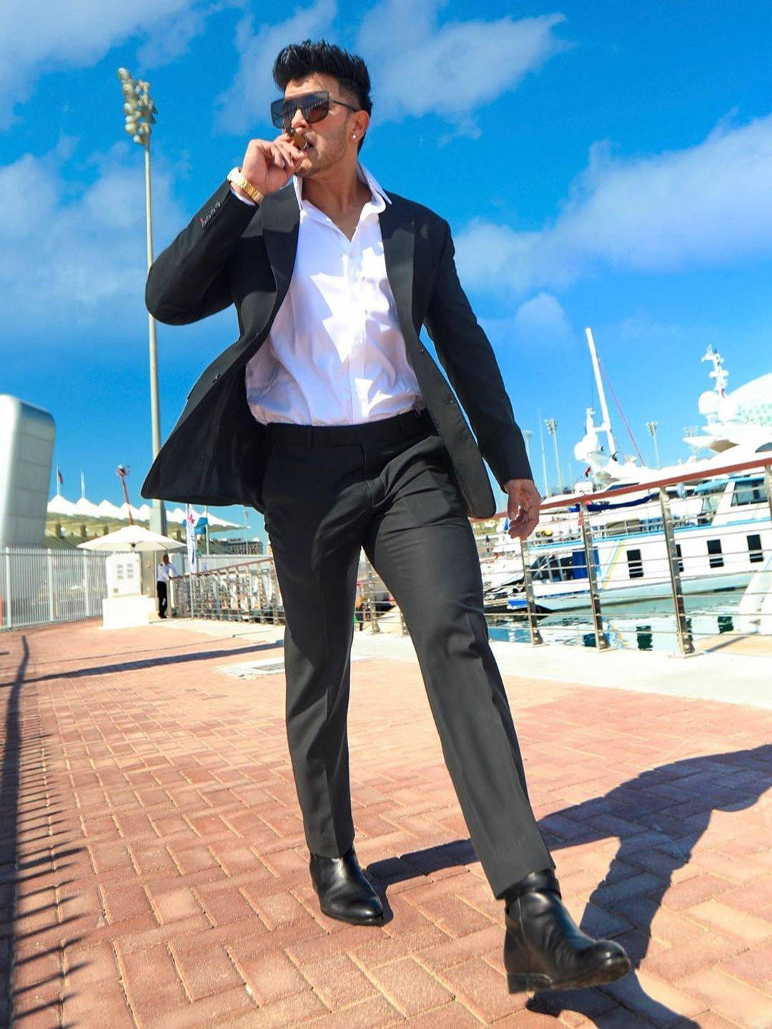 Dubai 桌布 壁紙 — 一位穿著黑色西裝和太陽眼鏡的時尚男士，在明亮的藍天下自信地走在豪華遊艇旁的磚砌碼頭上。