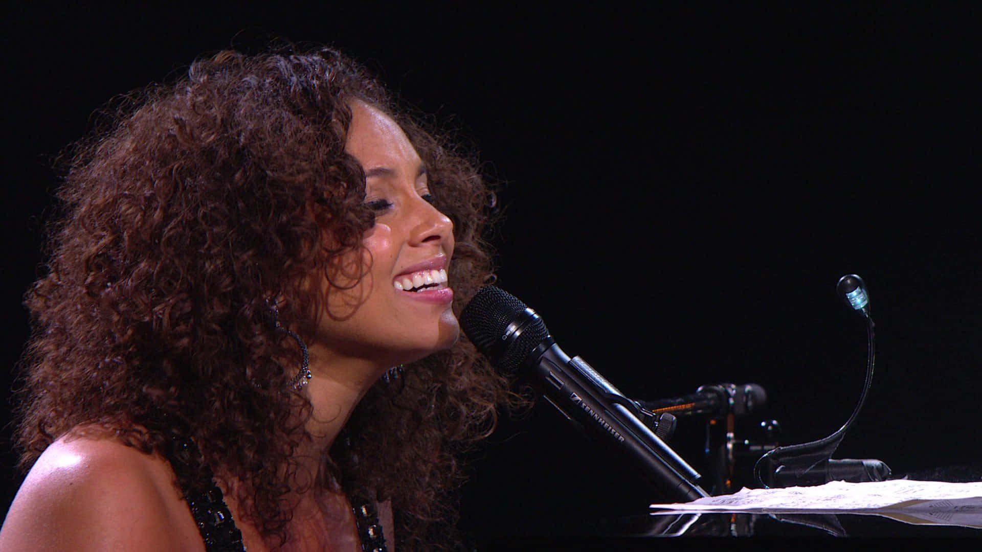 Alicia Keys 桌布 壁紙 — 一位笑容滿面、擁有豐盈曲髮的女性在黑暗的舞台上對著麥克風唱歌。