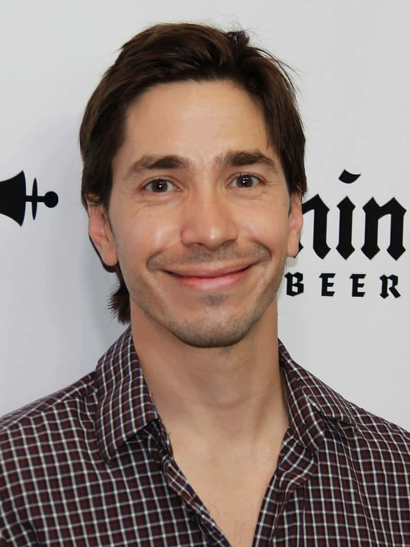 Justin Long 桌布 壁紙 — 一位笑容滿面、棕色頭髮、穿著格仔恤衫嘅男士企喺白色背景前，背景有黑色文字同標誌。