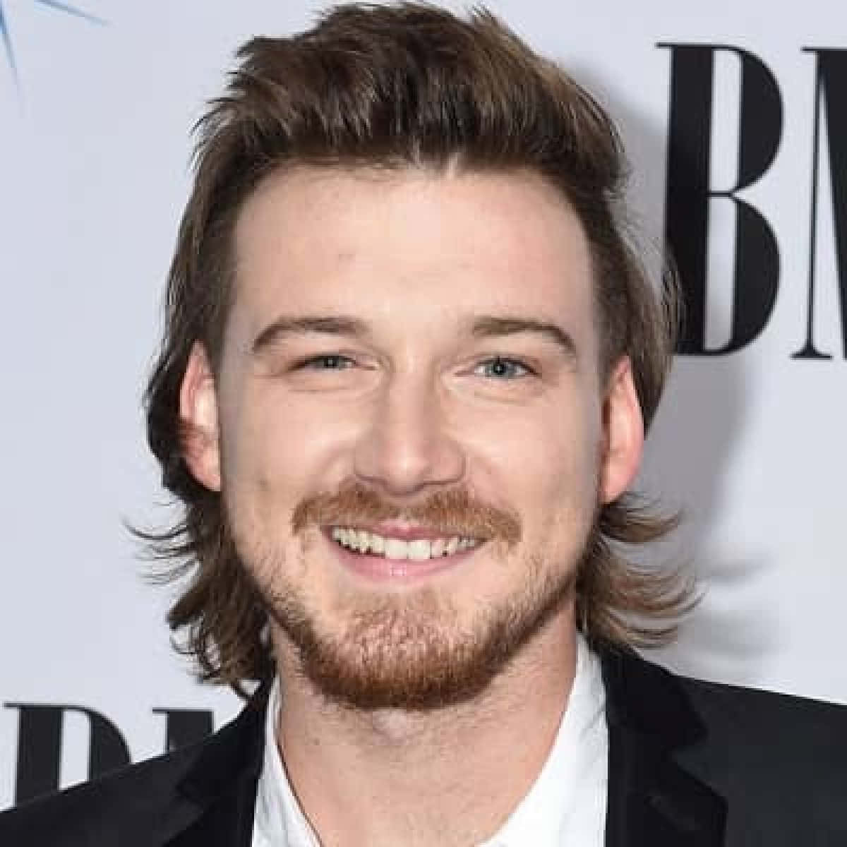 Morgan Wallen 桌布 壁紙 — 一位笑容滿面、棕色頭髮和鬍鬚的男士，身穿黑色西裝，背景為白色，帶有粗體字樣。