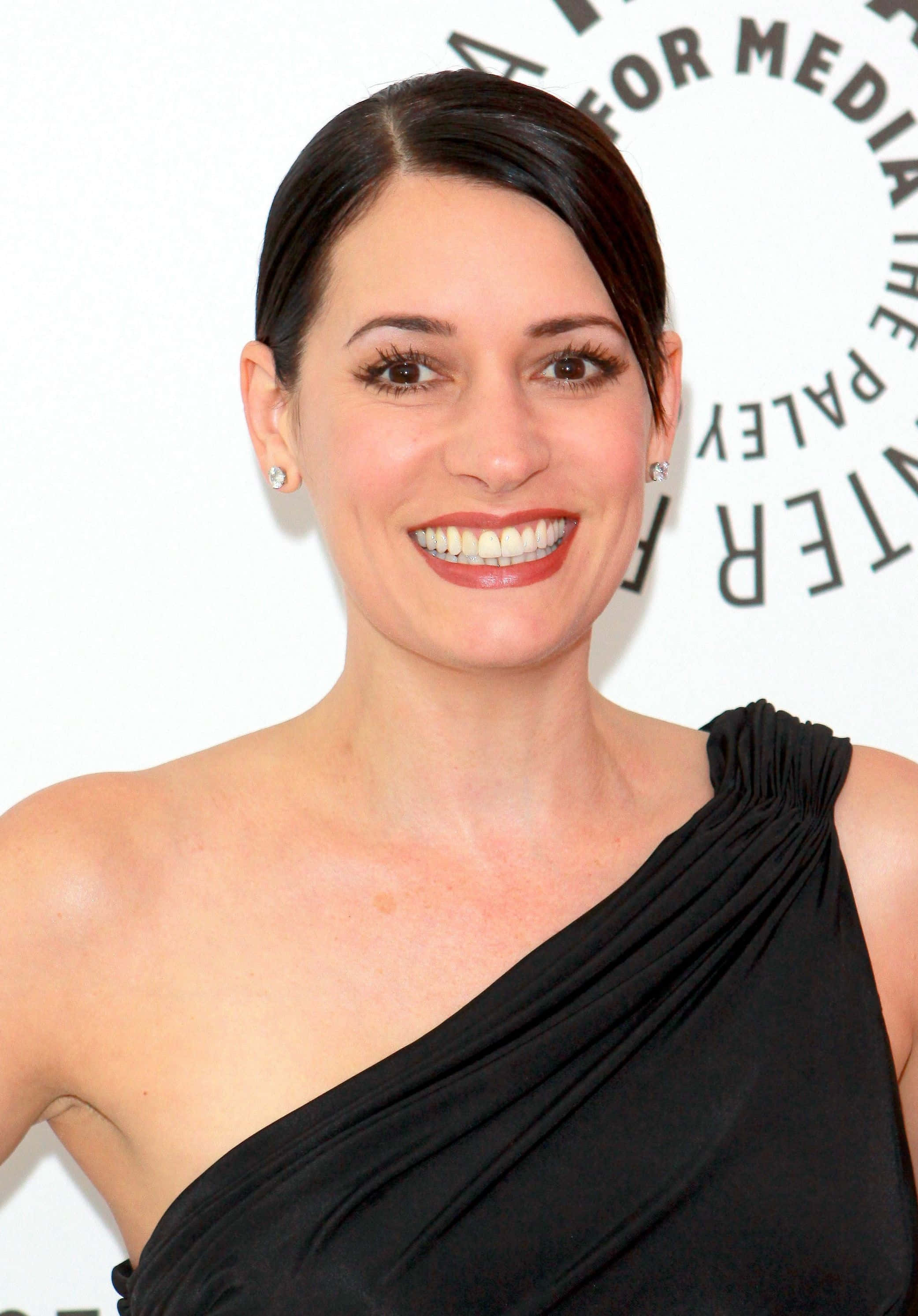 paget brewster 桌布 壁紙 — 一位笑容滿面、深色頭髮、塗著紅色唇膏的女性，在媒體活動中穿著黑色單肩連衣裙。