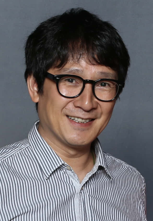 Ke Huy Quan 桌布 壁紙 — 一位笑容滿面、深色頭髮、戴黑色框眼鏡的男士，穿著條紋襯衫，背景是純灰色。