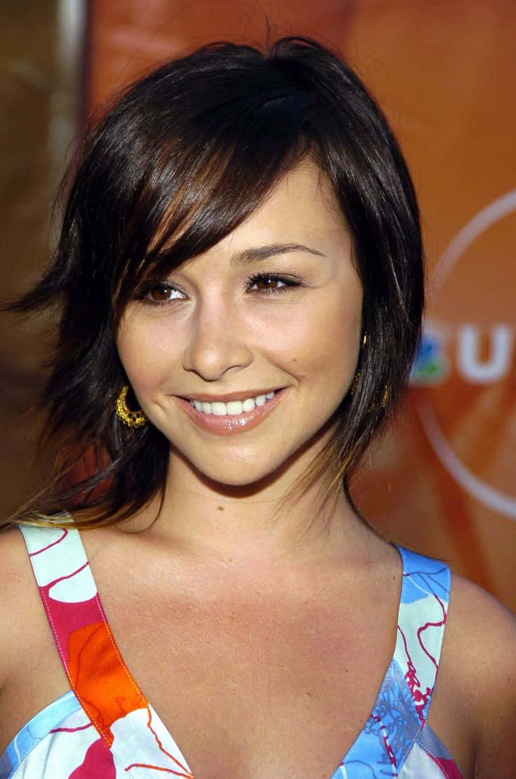 Danielle Harris 桌布 壁紙 — 一位笑容滿面、深色頭髮、穿著彩色圖案上衣的女性，在溫暖的橙色背景前擺姿勢。