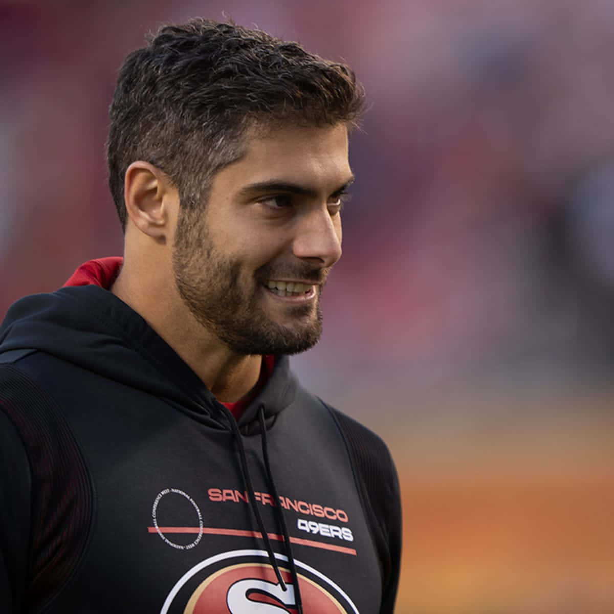 Jimmy Garoppolo 桌布 壁紙 — 一位笑容滿面、深色頭髮和鬍鬚的男子，在模糊的體育場背景下穿著黑色三藩市49人連帽衫。