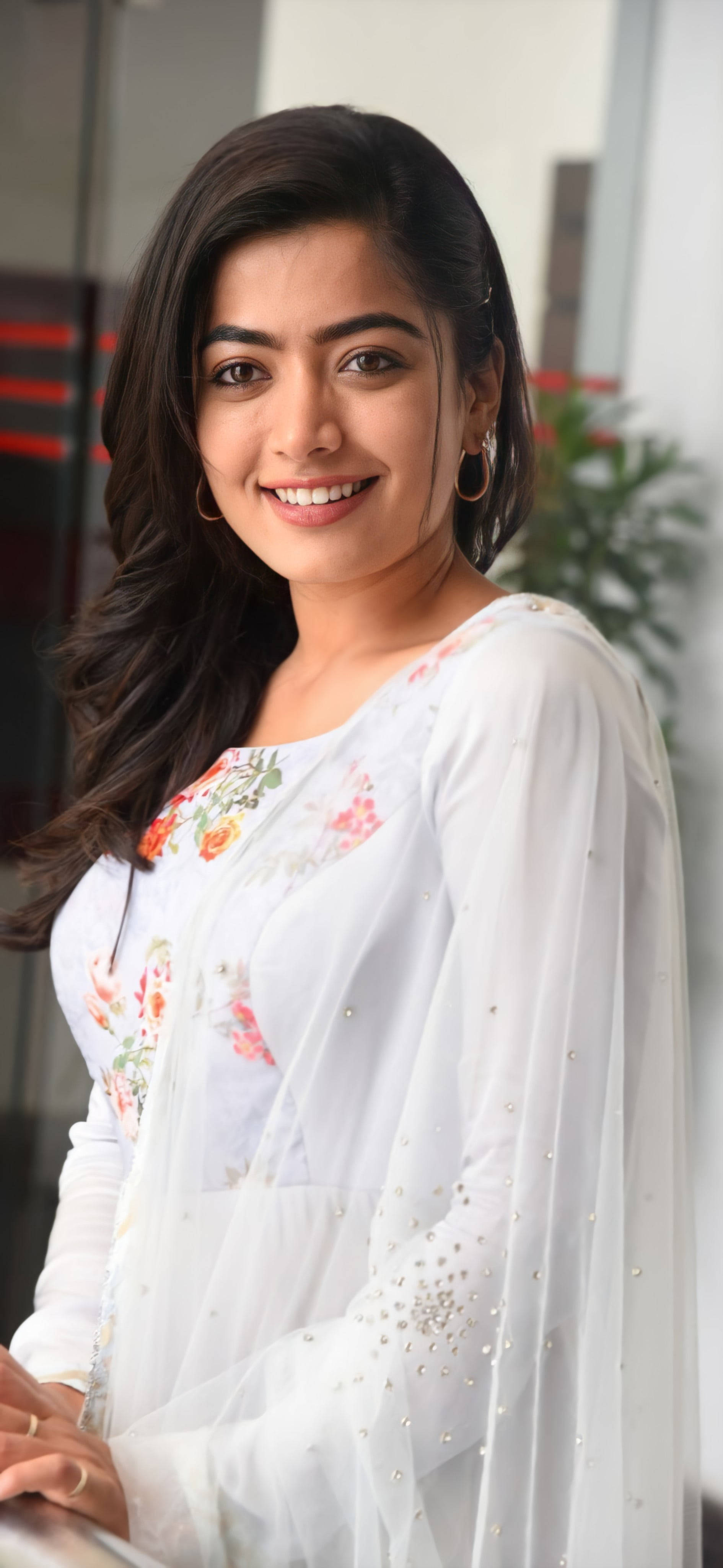 telugu 桌布 壁紙 — 一位笑容滿面、深色頭髮的女性，穿著白色花卉刺繡服裝，背景是現代室內環境。