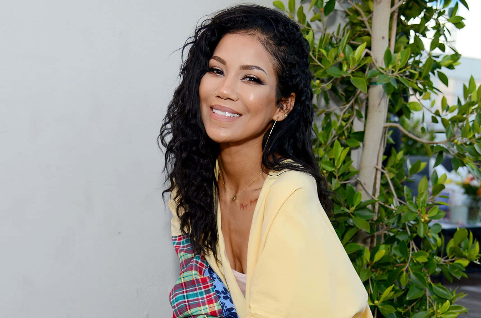 jhene aiko 桌布 壁紙 — 一位笑容滿面、留著深色長曲髮的女性，身穿黃色開襟毛衣和格仔上衣，背景是白色牆壁和綠色植物。
