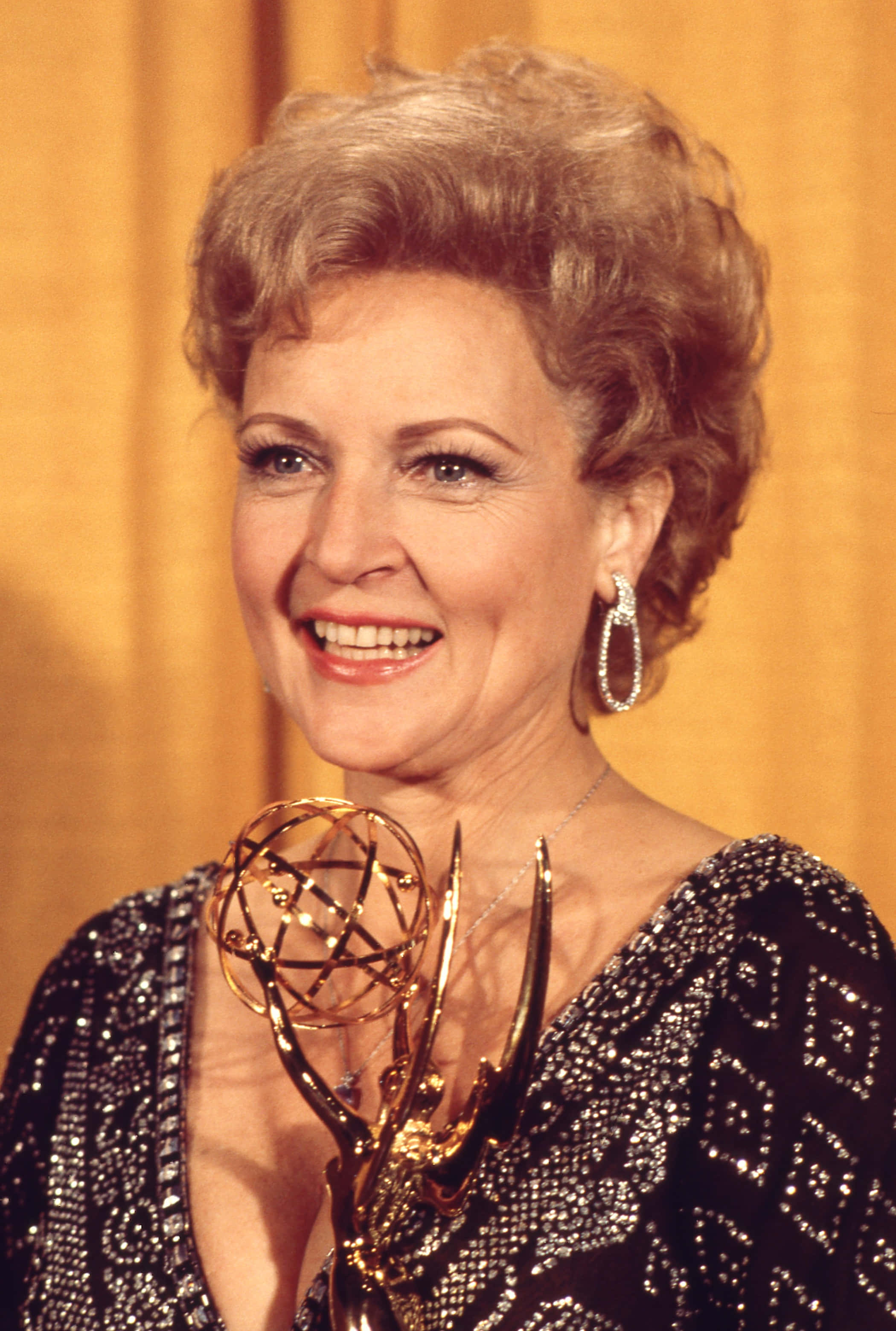 Betty White 桌布 壁紙 — 一位笑容滿面、頭髮造型時尚的女性，在溫暖的黃色窗簾背景下手持金色艾美獎獎座。