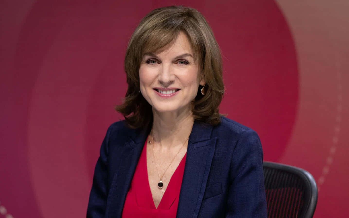 Fiona Bruce 桌布 壁紙 — 一位笑容滿面的棕髮女性，在鮮紅色圓形背景前，身穿海軍藍西裝外套，內搭紅色上衣。
