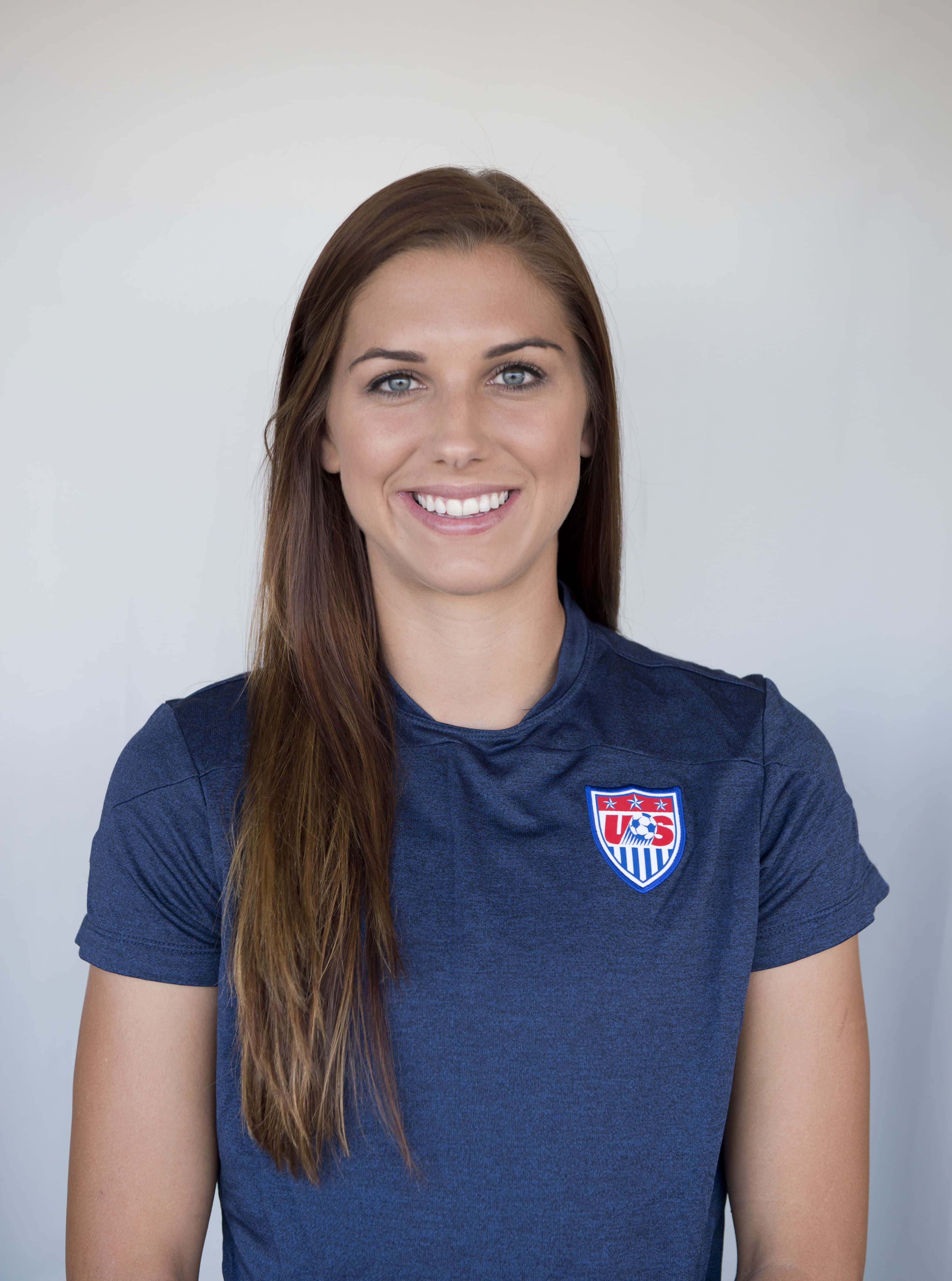 Alex Morgan 桌布 壁紙 — 一位笑容滿面的長髮棕髮女性，在純白背景前穿著深藍色美國足球球衣。