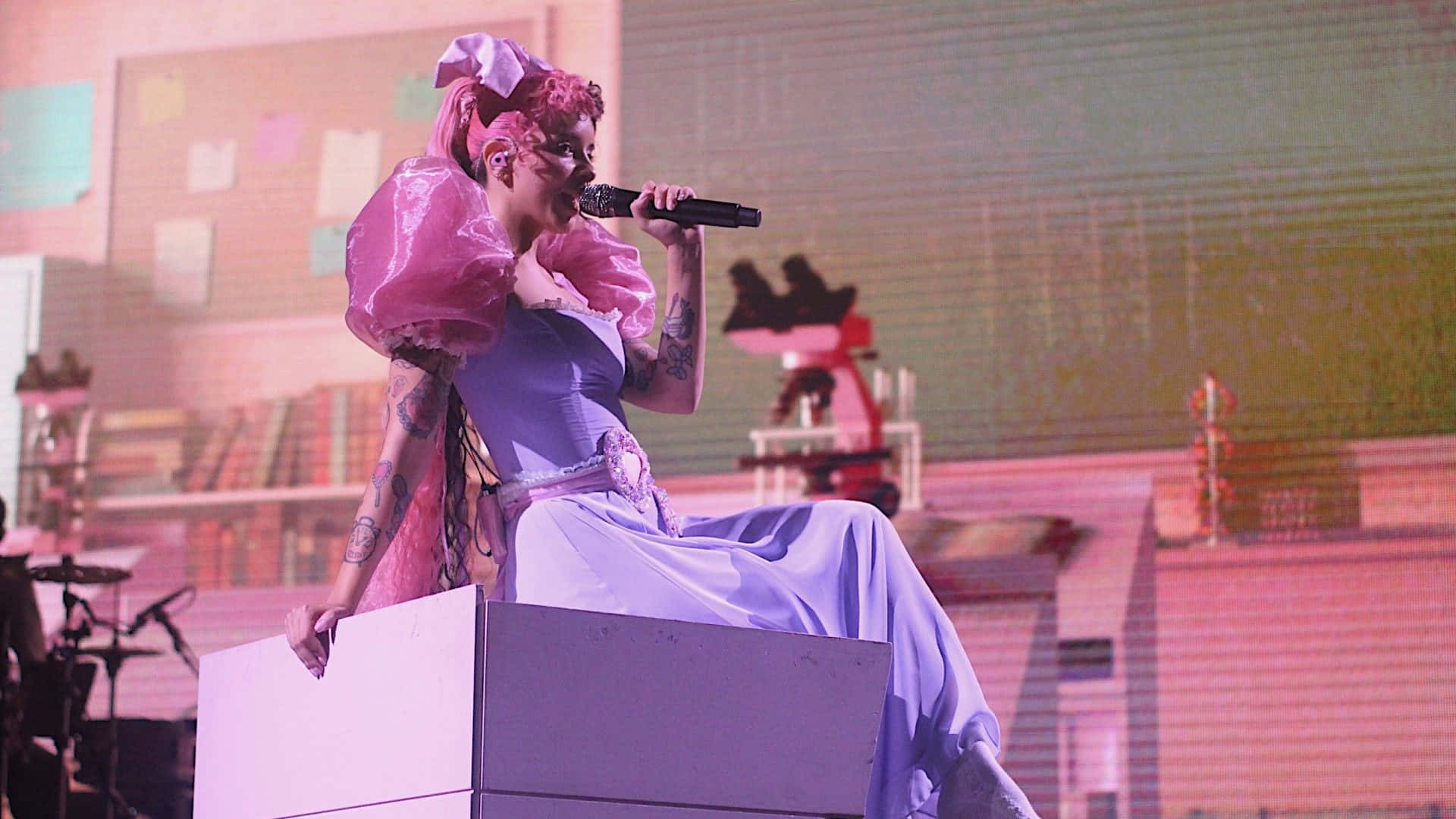 Melanie Martinez 桌布 壁紙 — 一位粉紅色頭髮、穿著泡泡袖的表演者坐在舞台上一個白色箱子上，在粉紅色燈光下對著咪高峰唱歌。