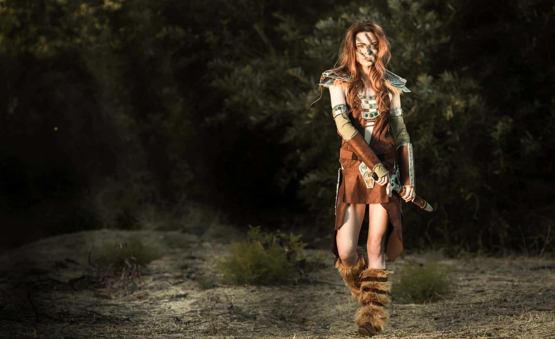 Aela The Huntress 桌布 壁紙 — 一位紅髮、臉上塗有部落圖騰的女戰士，在沐浴著金色陽光的森林空地中大步向前。