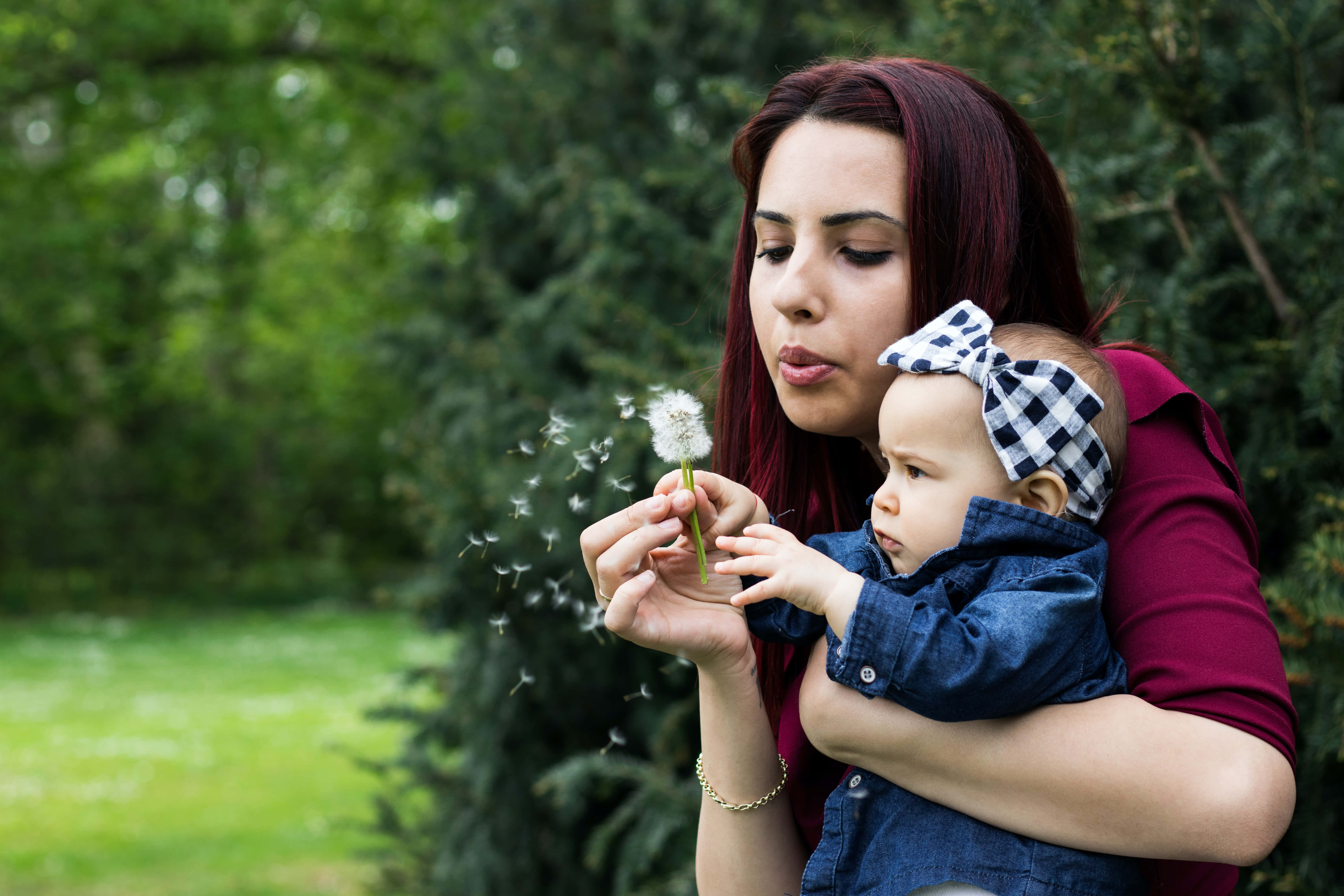 Mom 桌布 壁紙 — 一位紅髮女士在綠樹成蔭的公園裡抱著嬰兒，輕輕吹散蒲公英種子。