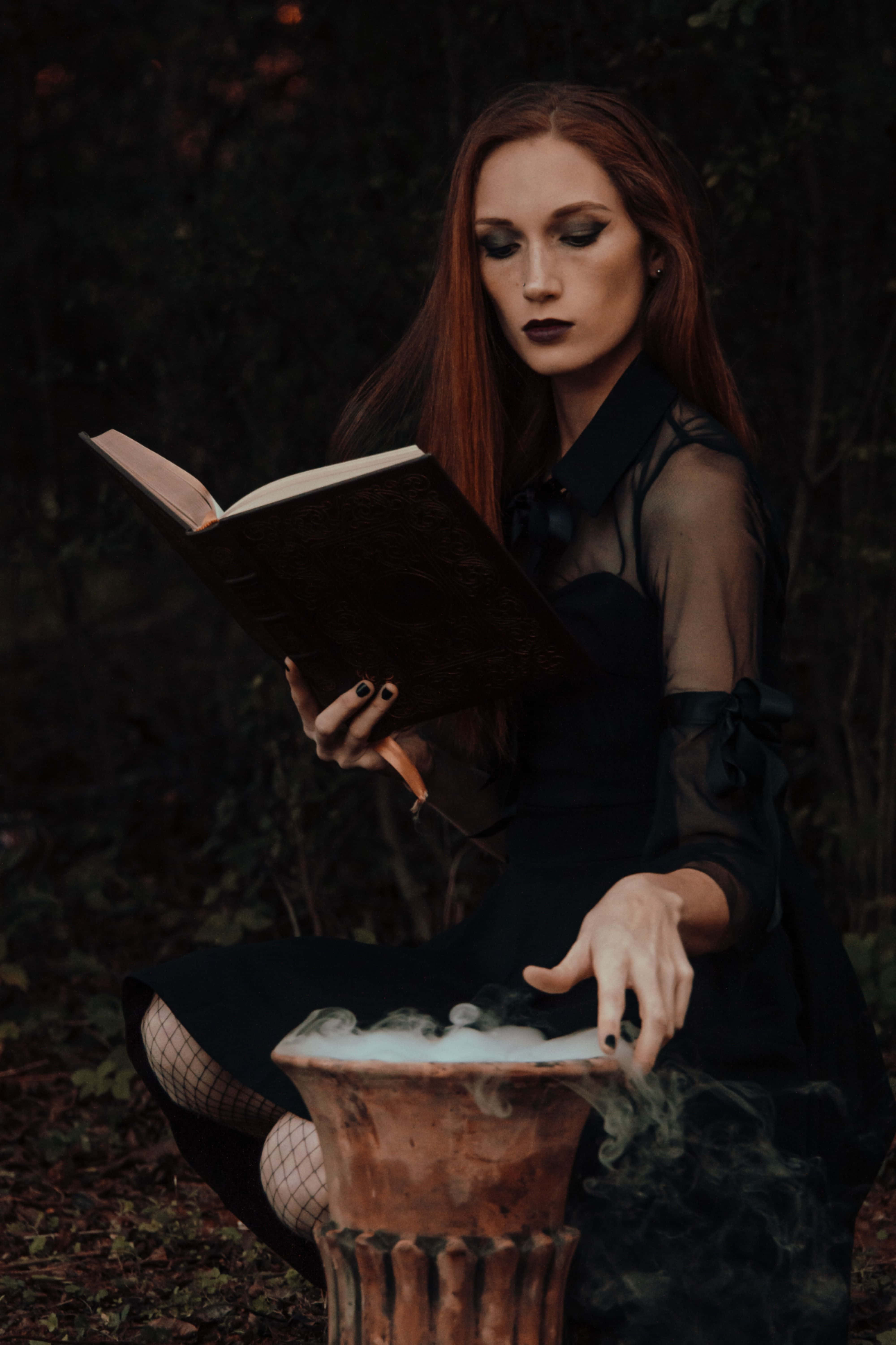 Witch Costumes 桌布 壁紙 — 一位紅髮女子在黑暗的森林中閱讀一本書，她的手懸在一個冒著蒸汽的大鍋上方。