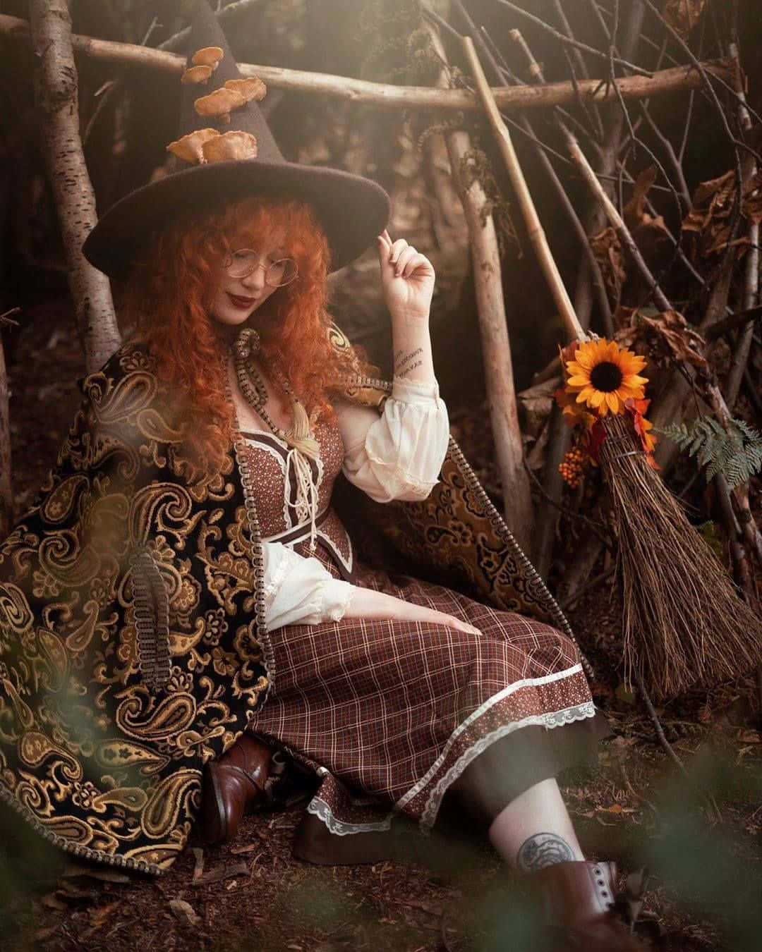 Witch Costumes 桌布 壁紙 — 一位紅髮女巫穿著華麗服飾，優雅地坐在陽光斑駁的森林中，她的帽子上裝飾著蘑菇，旁邊放著一把掃帚。