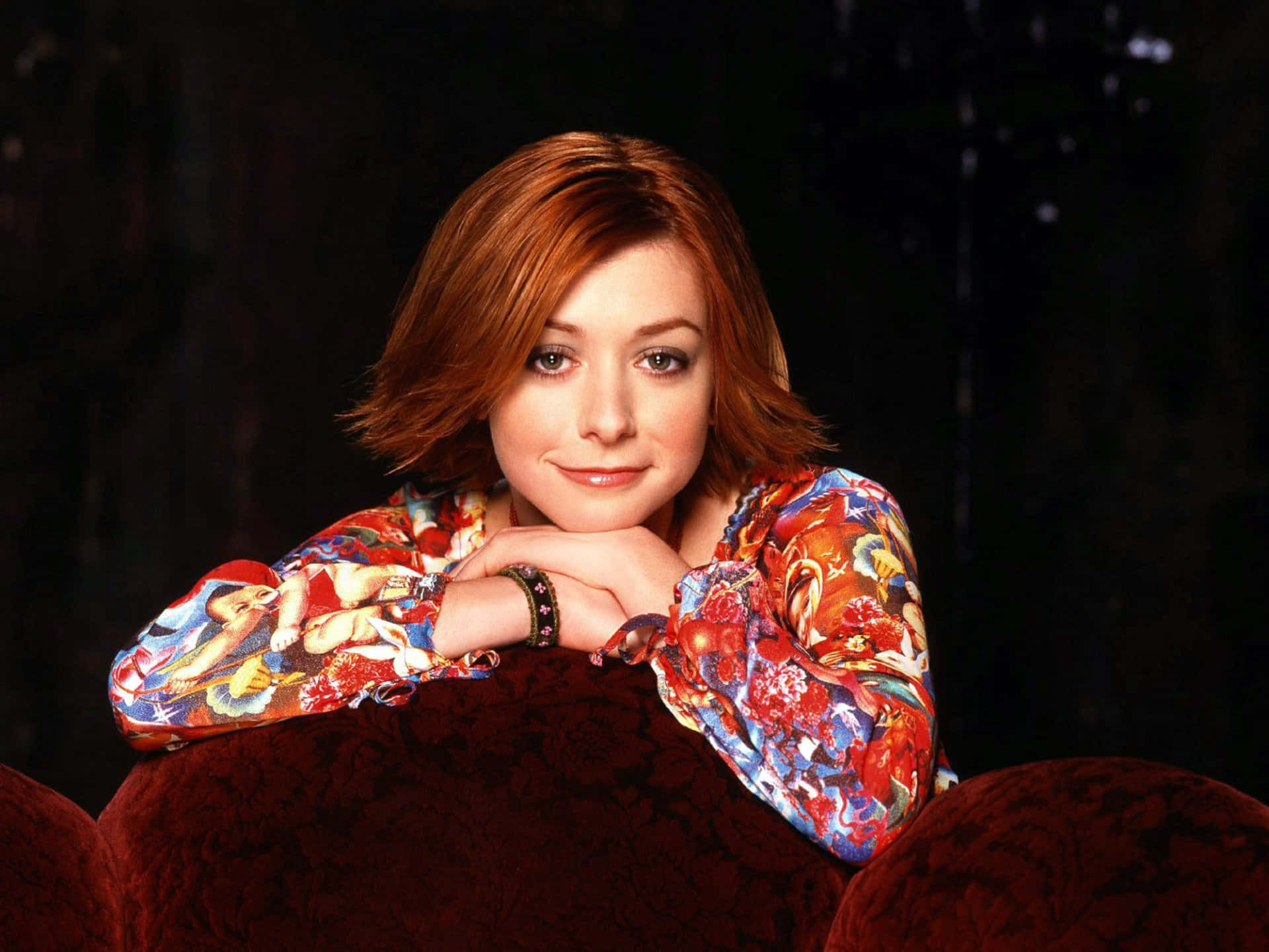 Alyson Hannigan 桌布 壁紙 — 一位紅髮女性在深色背景下穿著色彩繽紛的花卉襯衫，將下巴擱在手上。