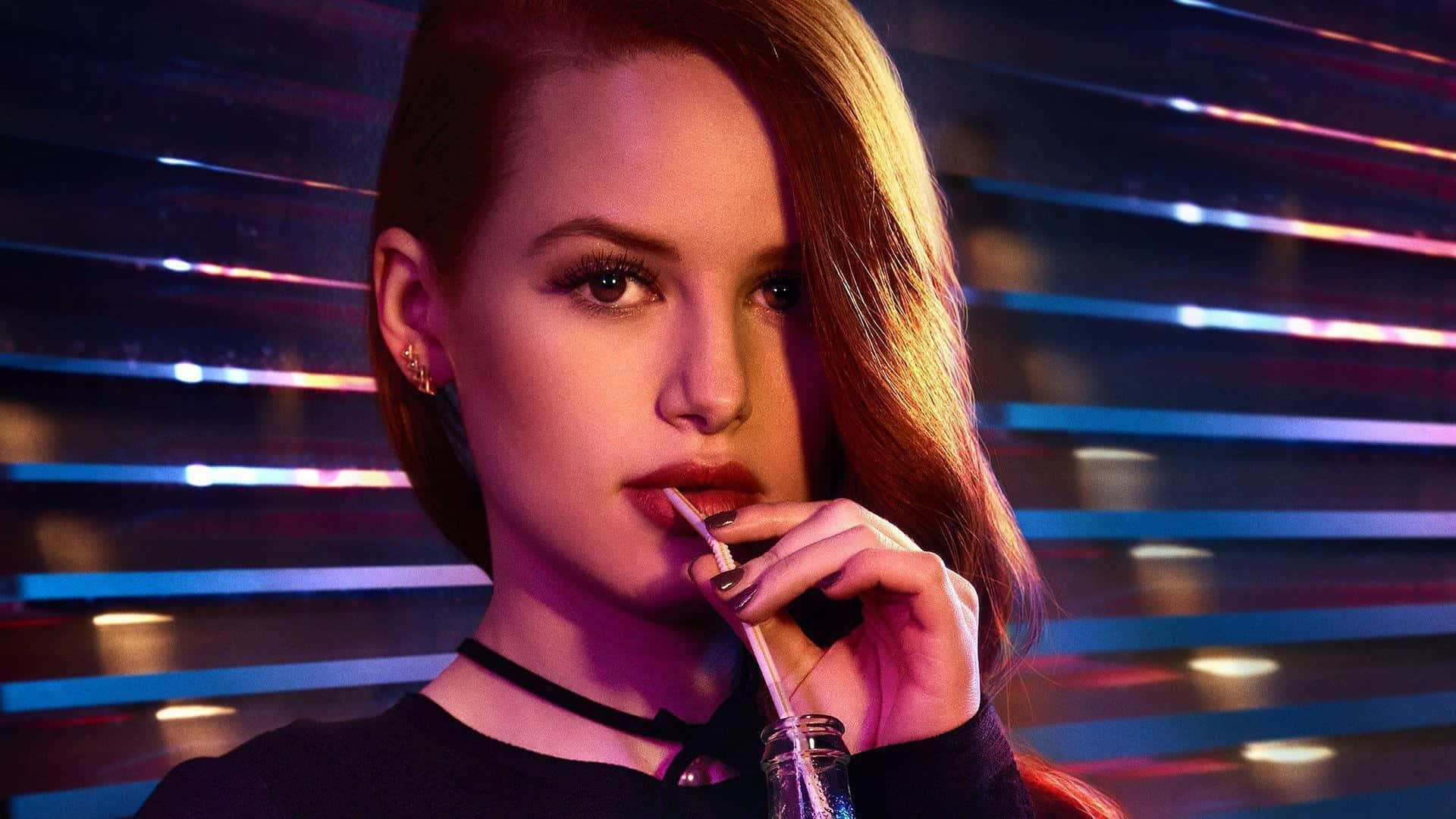 Riverdale 桌布 壁紙 — 一位紅髮女性用吸管啜飲玻璃杯中的飲料，背景是充滿活力的霓虹燈光。