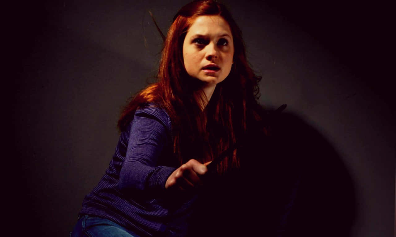 Ginny Weasley 桌布 壁紙 — 一位紅髮年輕女性在昏暗的攝影棚中，手持魔杖向前指，表情專注而強烈。