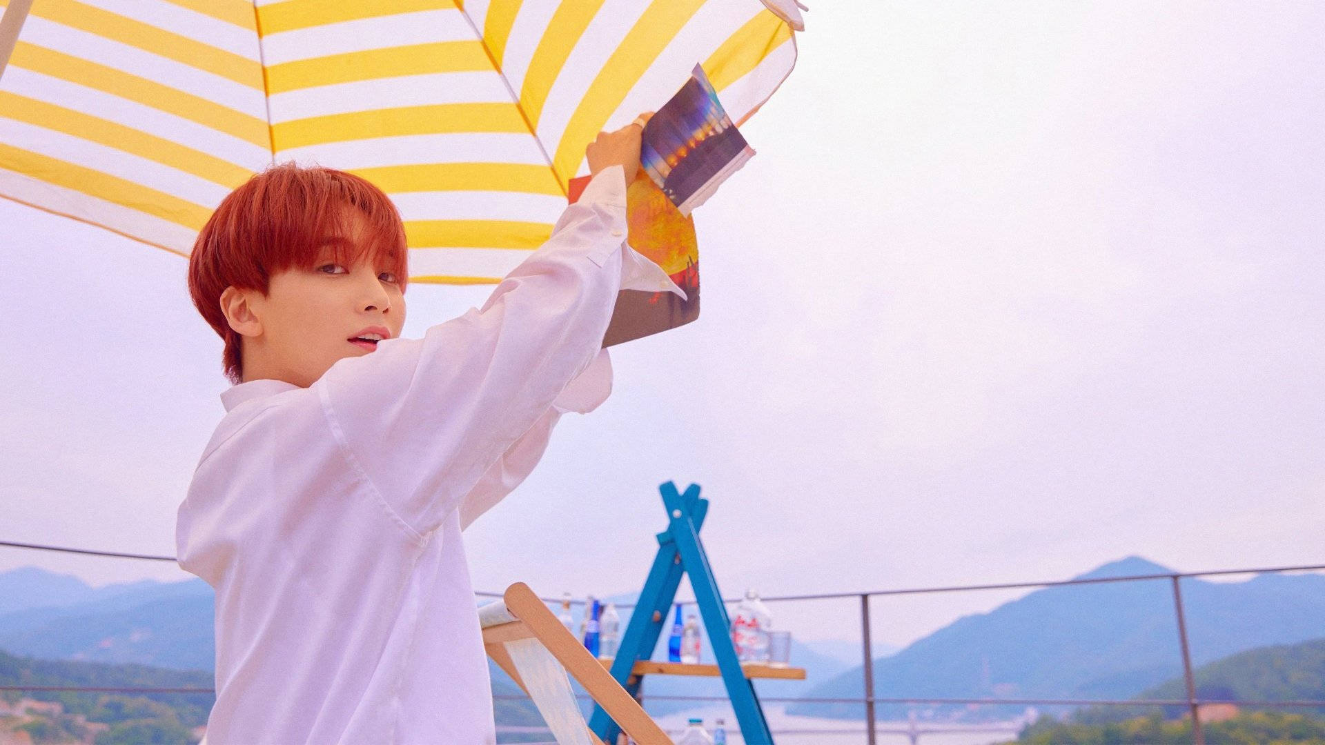 Yoon Jeonghan YMMDay 壁紙 — 一位紅髮年輕男子站在陽台上，俯瞰著柔和天空下的山脈，手持一把黃白條紋雨傘。