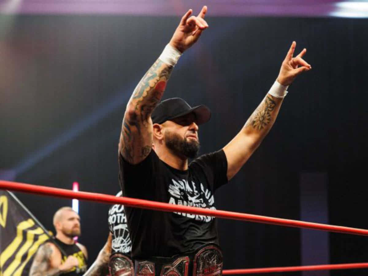 Rock And Roll Karl Anderson 壁紙 — 一位紋身摔角手身穿黑色襯衫和帽子，在紅色擂台上高舉雙手慶祝勝利，並持有冠軍腰帶。