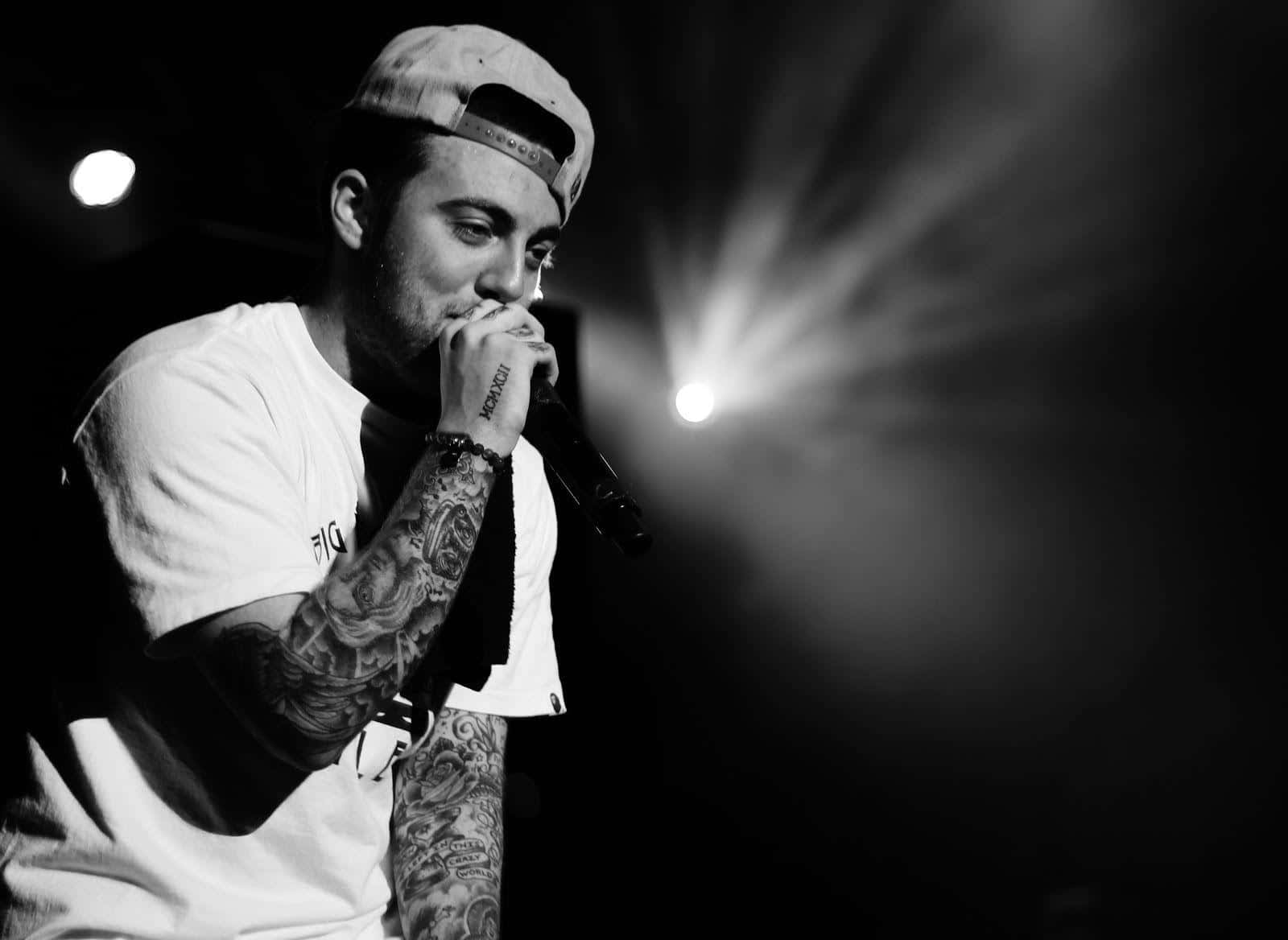 Mac Miller 桌布 壁紙 — 一位紋身音樂家戴著帽子，在戲劇性的舞台燈光下以黑白風格對著麥克風唱歌。