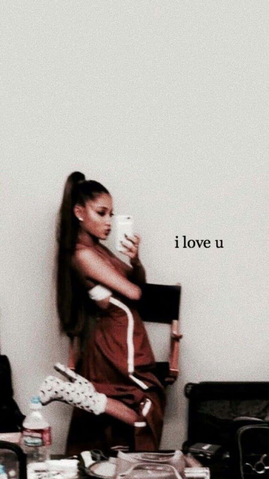 Aesthetic Ariana Grande 桌布 壁紙 — 一位紮著高馬尾的女性坐在椅子上，手持電話，身後牆上可見「i love u」字樣。