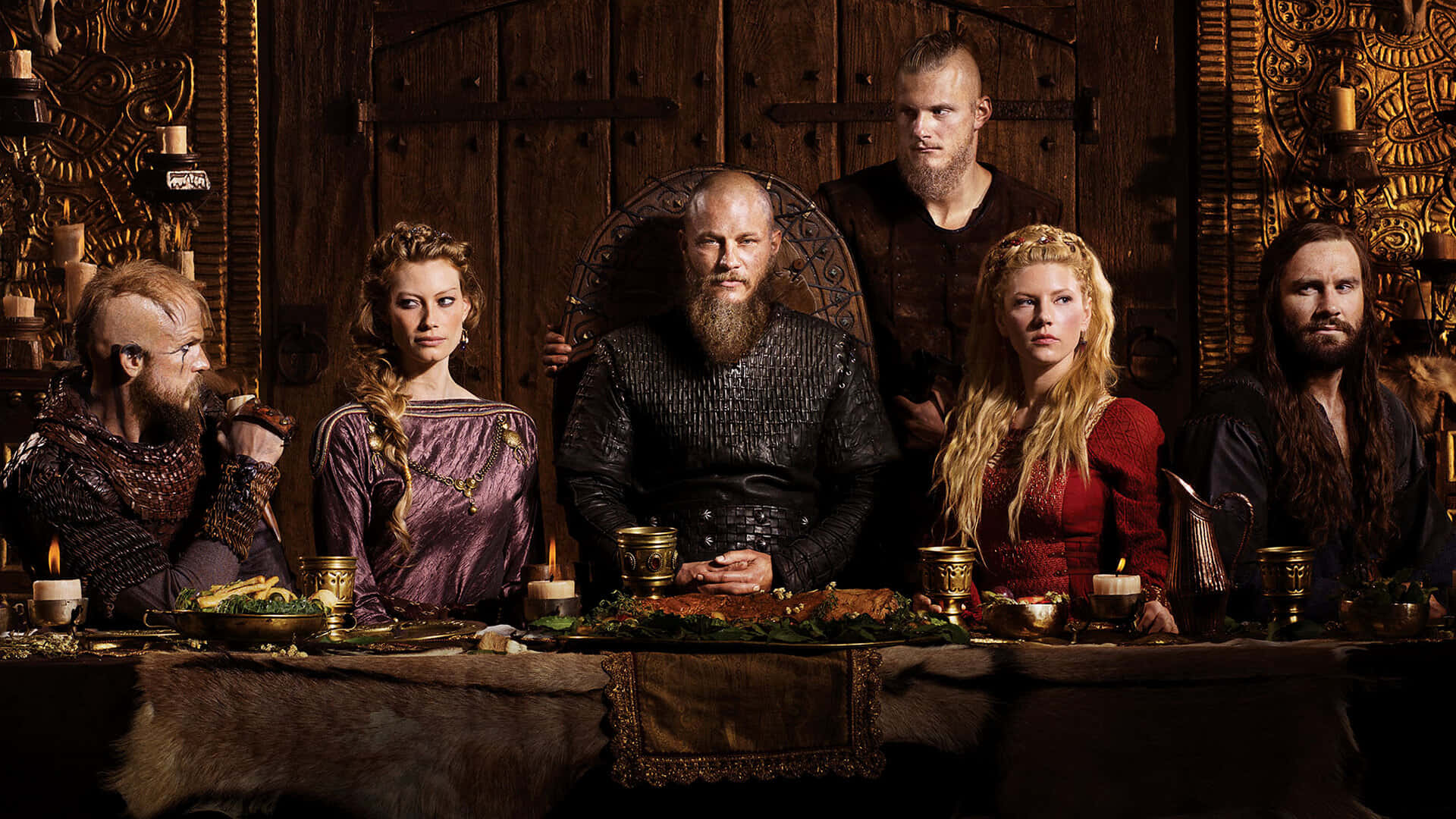 Vikings 桌布 壁紙 — 一位維京首領和他的船員莊嚴地坐在一個裝飾著複雜雕刻的木製大廳裡，參加一場燭光盛宴。