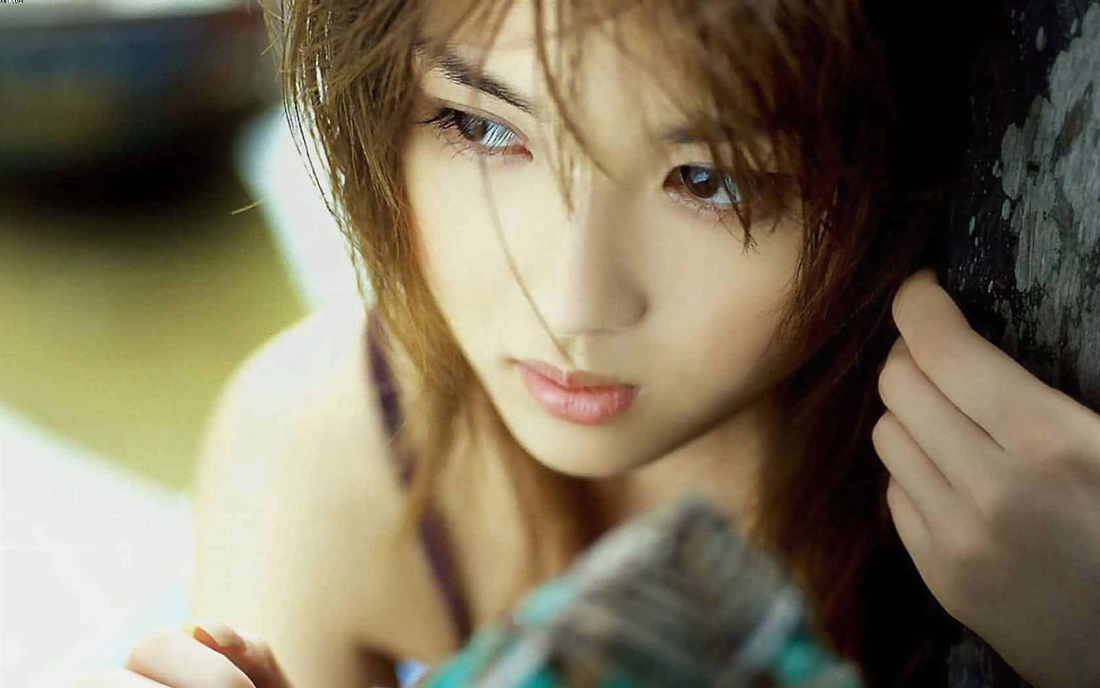 一位美麗女孩自拍 壁紙 — 一位年輕女性，一頭凌亂的棕色頭髮，在溫暖的環境光線中靠著紋理牆壁，溫柔地凝視著。