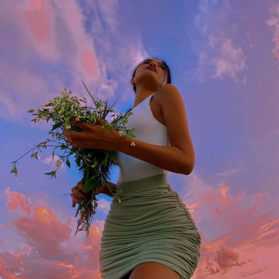一位美麗的美學女孩凝望夕陽 壁紙 — 一位身穿白色上衣和綠色裙子的女士，在夢幻的粉紫色日落天空下捧著野花。