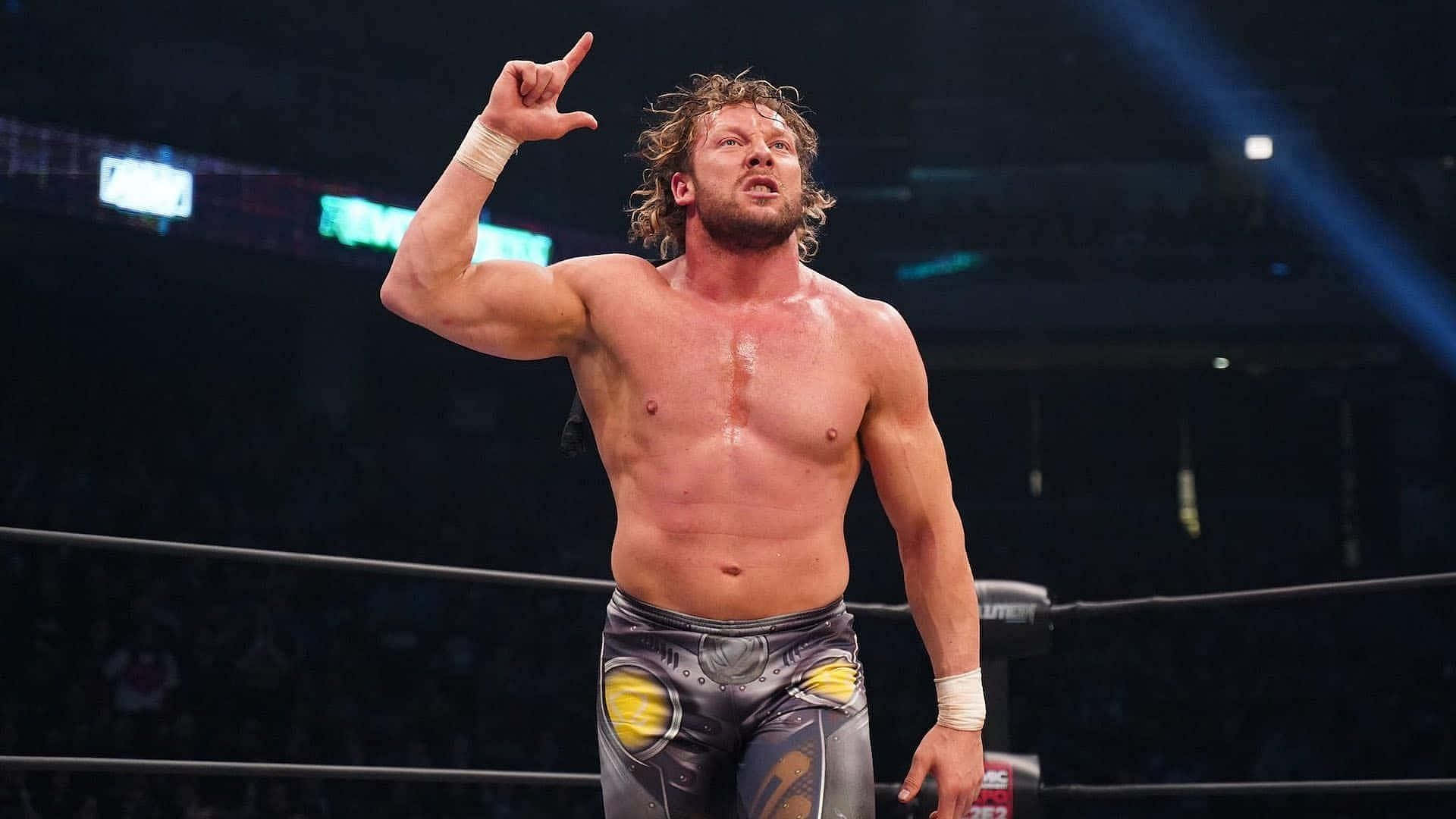 Kenny Omega Gray And Yellow Pants 壁紙 — 一位肌肉發達、捲髮的摔角手在明亮的場館燈光下於擂台上指向上方，背景有群眾。