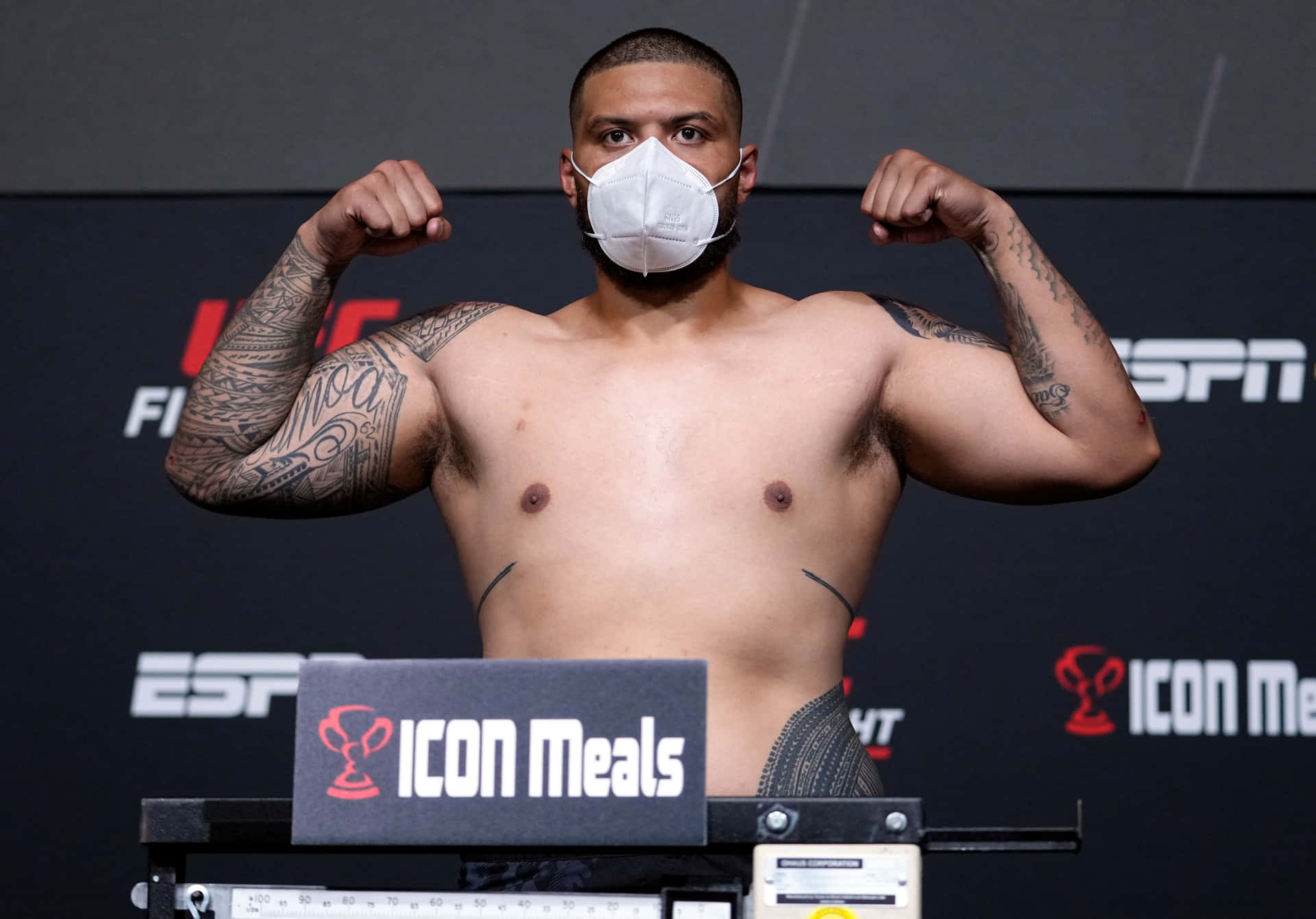 UFC 桌布 壁紙 — 一位肌肉發達、有紋身的拳手在UFC稱重活動中戴著白色口罩彎曲手臂。